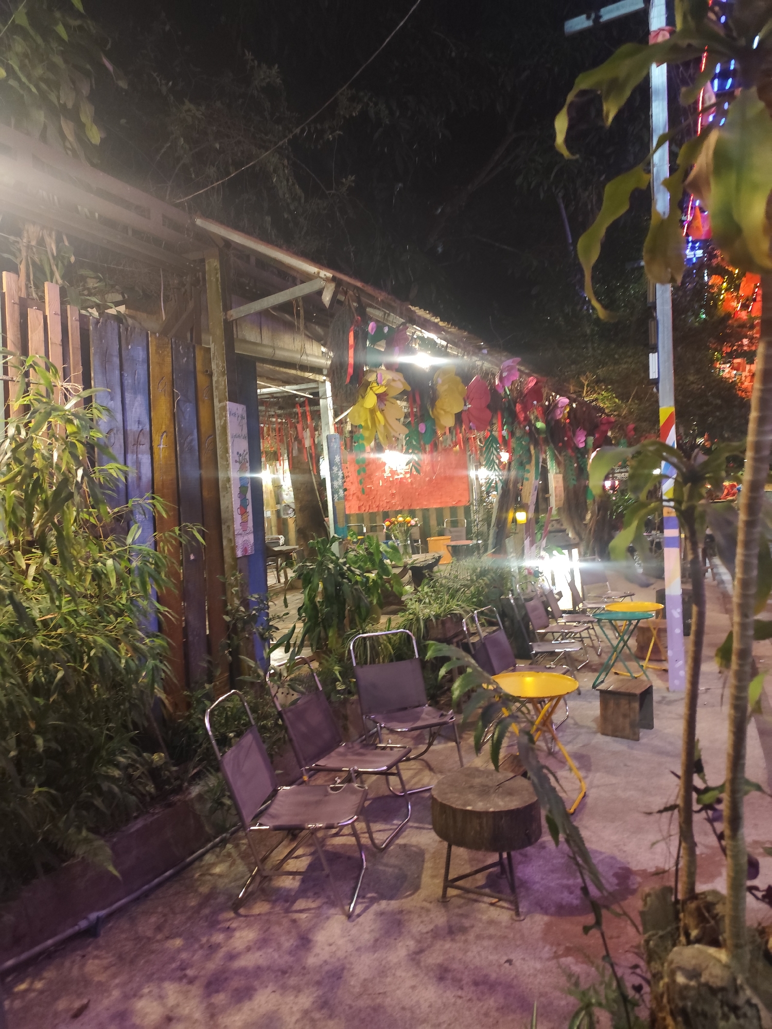 The image depicts an outdoor seating area, likely in Da Lat, Vietnam, at night. Several metal folding chairs with dark purple fabric seats are arranged around small, colorful tables - some yellow, some green - and tree stump-like stools. Lush greenery surrounds the seating area, with plants flanking the entrance on the left and growing along the ground. Overhead, the space is decorated with colorful, hanging decorations. The building on the left is constructed of vertical wooden planks, some painted in shades of blue and yellow. The scene is illuminated by artificial light sources, creating a warm ambiance. The foreground is filled with a concrete pathway, and the background fades into the darkness, revealing trees and more decorations. No people are visible in the image.