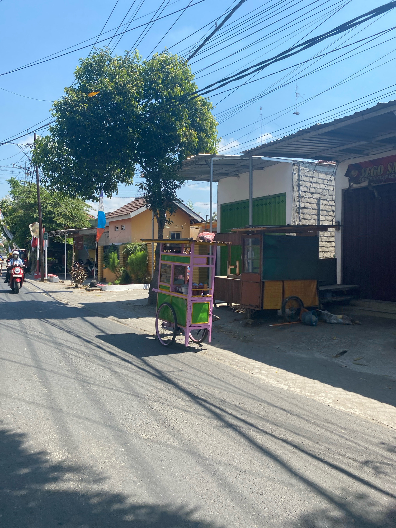 The image depicts a street scene in the city of Kediri, Indonesia, on a clear day. A red scooter with a rider wearing a white helmet is traveling on the asphalt road from left to right. Beside the road, a brightly colored food cart, predominantly pink and green, is parked. Behind the food cart, a longer, brown food stall with a wooden structure and metal roofing is also situated. The stall has a dark green panel and appears to be closed. Further down the street, a building with a white brick wall and a dark brown shuttered storefront is visible. A sign above this storefront reads "SEGO SA". Several overhead power lines crisscross the sky. Trees with lush green foliage are present on the left side of the image, casting shadows on the road, which is also partially covered by the shadows of the power lines. The overall environment suggests a typical street in a residential or commercial area of Kediri.