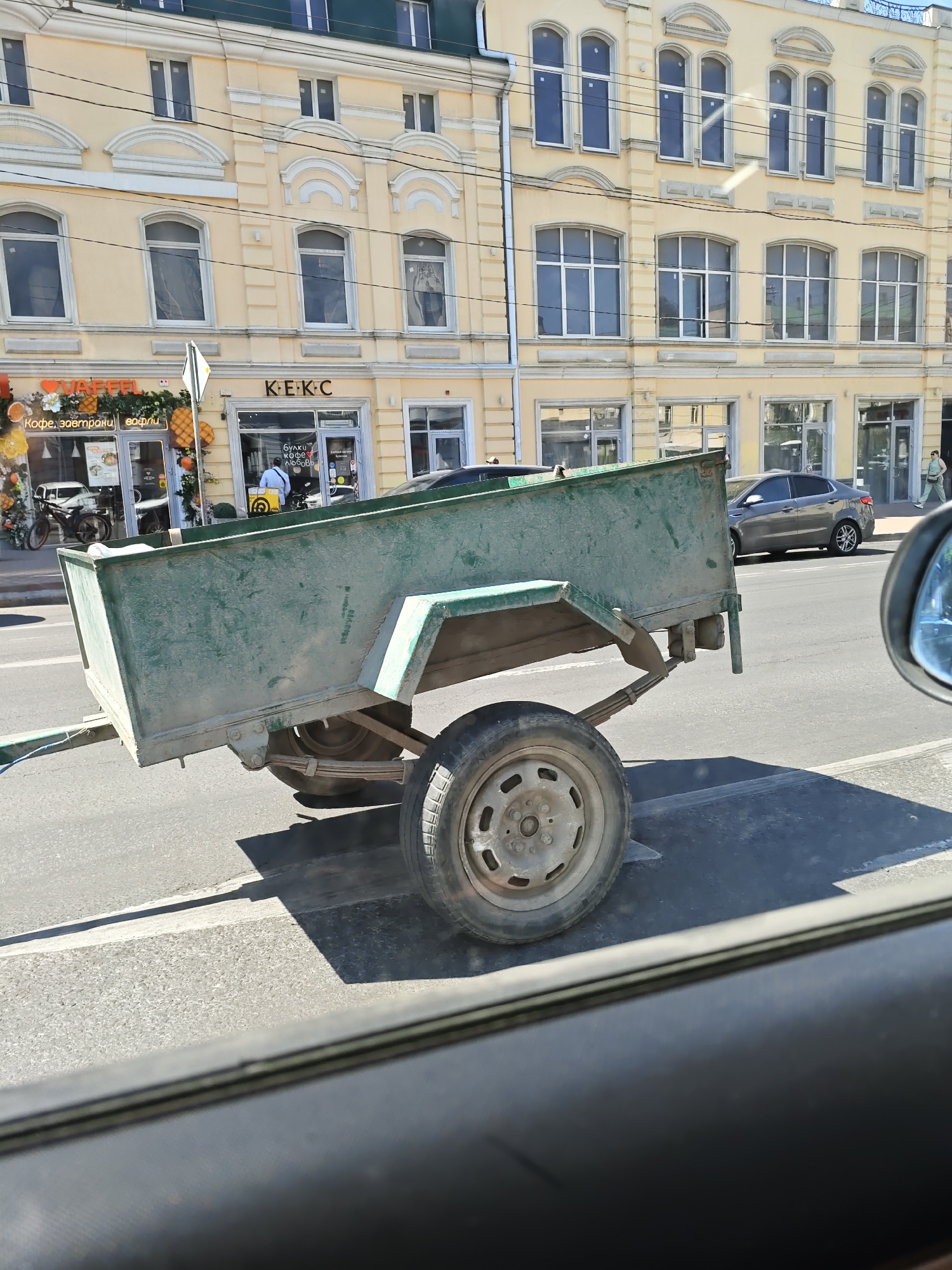 The image shows a green, dusty trailer parked on a paved road in the city of Rostov-on-Don, Russia. The trailer is the primary subject, taking up a significant portion of the frame. It appears to be a single-axle trailer with a metal body and visible leaf springs. A single wheel with a grey rim is visible on the right side of the trailer. In the background, a light-colored building with multiple windows and architectural details is visible. The building has signs indicating businesses, including "VAFFEL" and "K-E-K-C." A gray car is parked on the road in front of the building, and a person is walking on the sidewalk. The scene is lit by sunlight, casting shadows on the road and pavement. The perspective suggests the photo was taken from inside another vehicle.