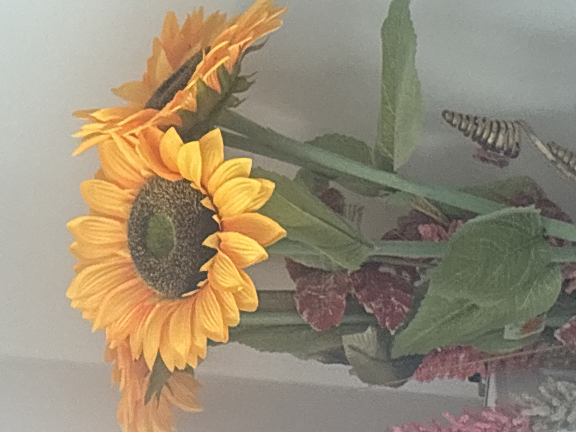 The image shows a close-up of a sunflower arrangement. The flowers are primarily yellow with dark brown centers. Green stems and leaves are visible behind the flowers. The background appears to be a light, neutral color.
There is no way to tell that this is from Strazhitsa, Bulgaria.