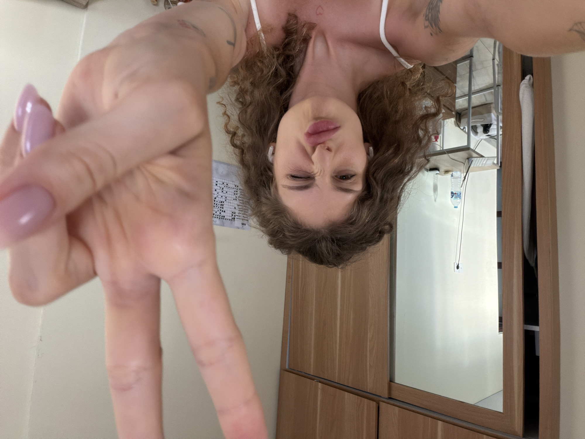 A woman with curly hair is taking a selfie. She is winking and holding up a peace sign with her left hand. She is wearing a white tank top and has tattoos on her arms and chest. The background includes a closet with mirrored doors, a desk, and a calendar. The image was taken at an upside-down angle. The location is not identifiable from the image. The city of North Kuta, Indonesia, is not identifiable from the image.