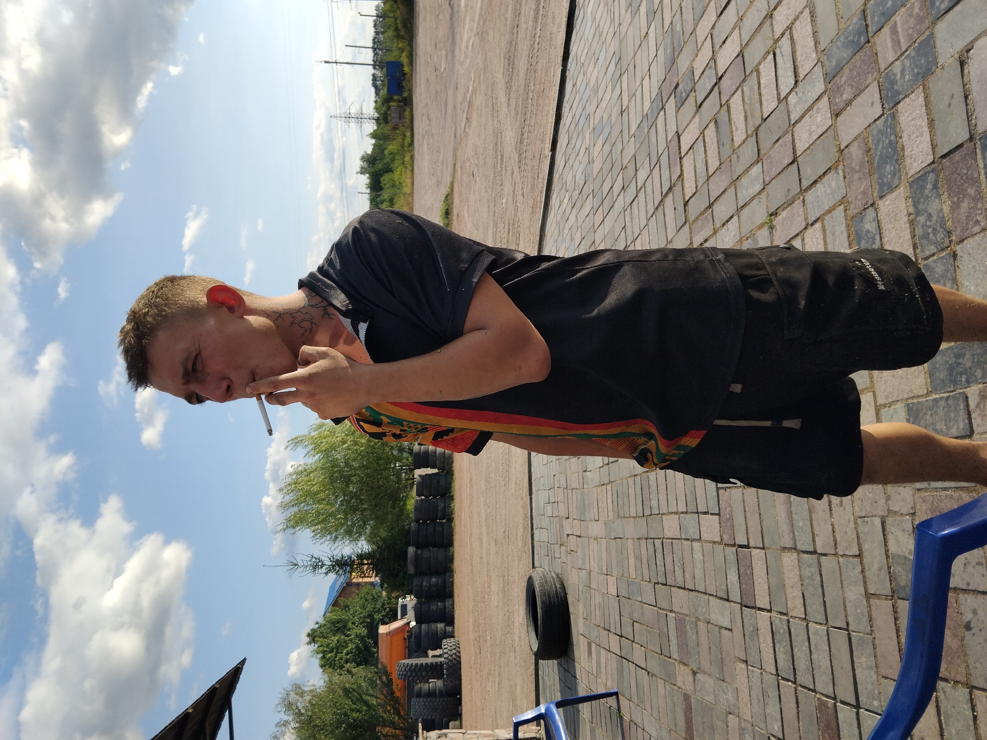 In Novoselytsia, Ukraine, a young man with short, light brown hair stands outdoors on a paved area with brick-like stones. He is wearing a black polo shirt with colorful accents on the sleeve and black cargo shorts. The man is smoking a cigarette, holding it to his mouth with his right hand. He has tattoos on his neck. In the background, there is a stack of tires, some trees, a building, and power lines against a blue sky with scattered clouds. A blue plastic chair is partially visible in the lower left corner of the image.