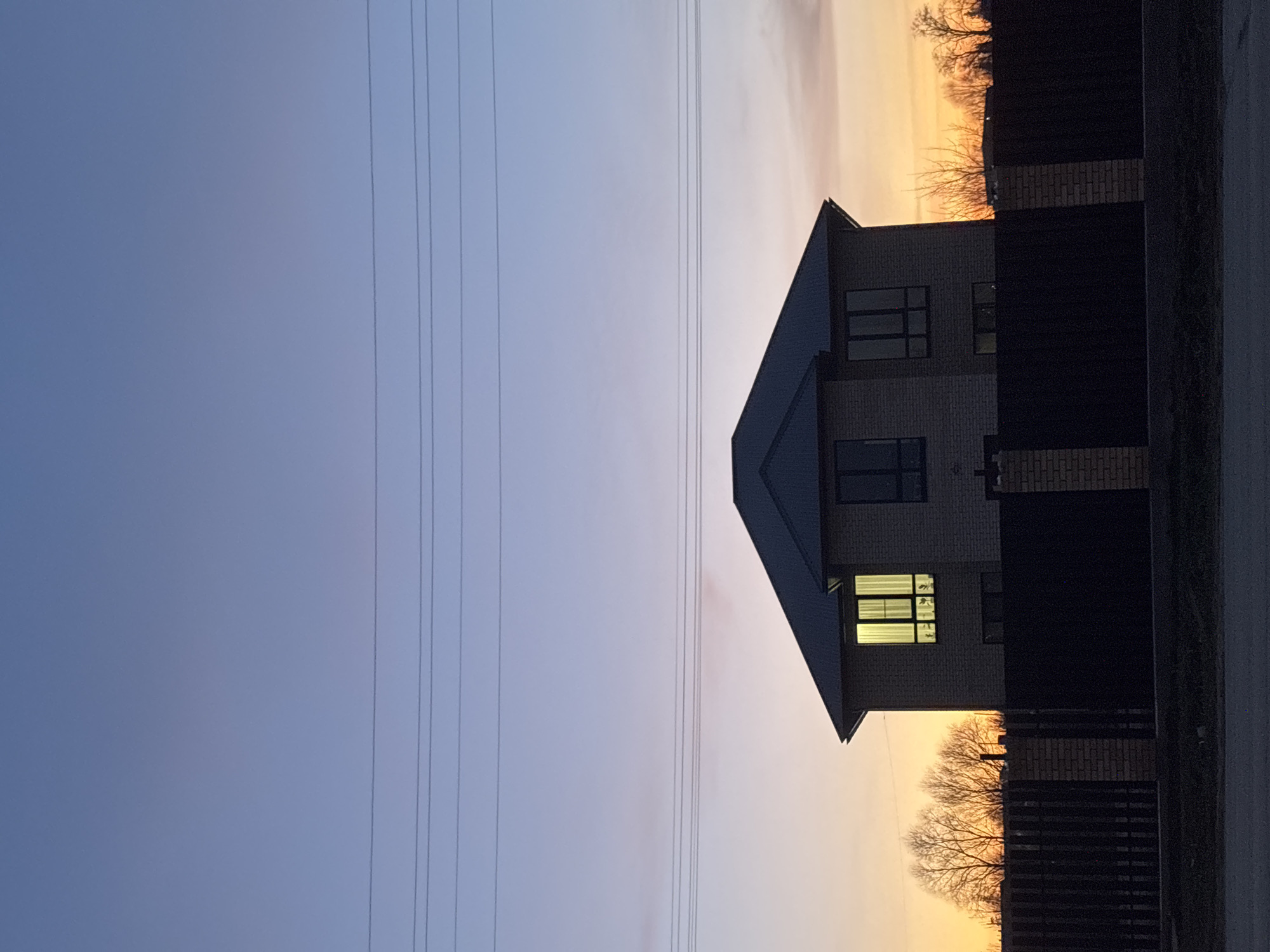 The image, taken in Фоки, Russia, depicts a residential scene during either sunrise or sunset. A two-story house with light-colored brick walls and a dark, sloped roof is positioned on the right side of the frame. Several windows are visible on the house; most are dark, reflecting the twilight sky, but one window on the lower floor emits a warm, bright light from within.
The sky displays a striking gradient, transitioning from a deep blue-purple at the top to a warm orange and yellow hue near the horizon, indicative of dawn or dusk. Multiple thin power lines stretch horizontally across the upper and middle sections of the sky. To the right of the house, a dark fence, likely made of metal or wood, runs parallel to the edge of the frame, partially obscuring the lower part of the horizon. Beyond the fence and house, the silhouettes of bare-branched trees are visible against the vibrant orange sky. A dark strip along the very bottom right suggests a road or ground. No people or visible text are present in the image, and the weather appears clear. The overall mood is serene and peaceful, capturing a quiet moment in a residential setting.