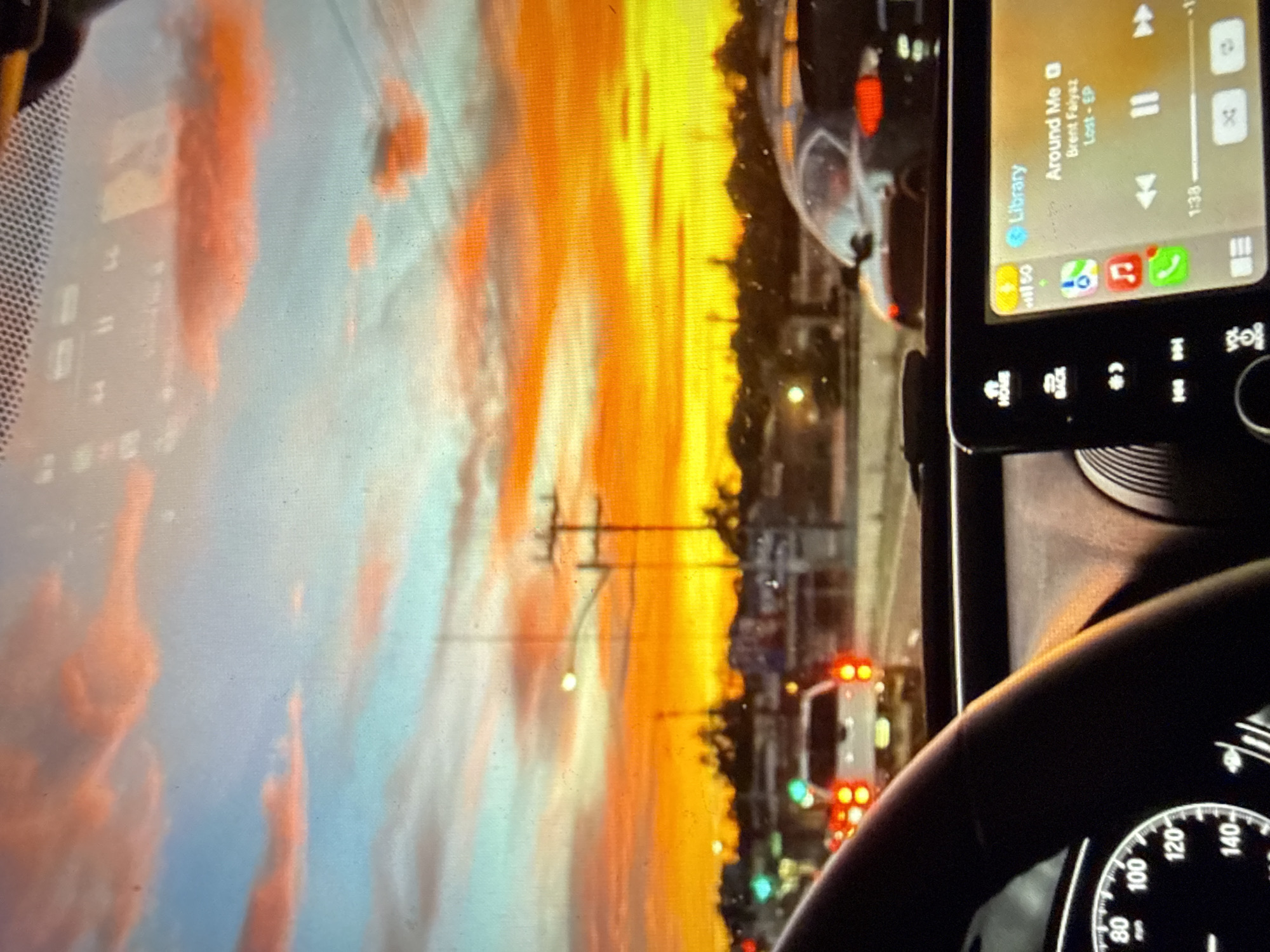 The image presents a first-person perspective from within a vehicle, looking through the windshield onto a roadway at sunset in the city of Kardzhali, Bulgaria. The sky is dominated by a dramatic sunset, transitioning from a pale blue at the top to vibrant orange and deep red, culminating in an intense yellow-orange glow above the horizon. Scattered clouds, illuminated from below, are visible across the upper portion of the sky.
In the midground, a multi-lane road is depicted with a moderate level of traffic. Utility poles, streetlights, and silhouetted structures and trees are visible along the distant horizon. Traffic lights, displaying both green and red signals, are present on the left side of the road. A white pickup truck with illuminated red taillights is positioned directly ahead in traffic. To the right of the pickup, a dark-colored sedan, potentially a Porsche, is partially visible, also displaying an illuminated red taillight.
The foreground shows the interior of the vehicle. A portion of a black steering wheel is visible on the left, partially obscuring the instrument cluster which includes a speedometer dial marked up to 140 (likely kilometers per hour). On the right, the vehicle's infotainment system screen is prominently displayed. The screen indicates a "Library" interface playing music, specifically the song "Around Me" by "Brent Faiyaz" from the album "Lost - EP," with a playback time of 1:38. Icons for cellular signal strength, battery level, and various applications such as phone, music, and navigation are visible on the screen. No individuals are explicitly visible within the frame.