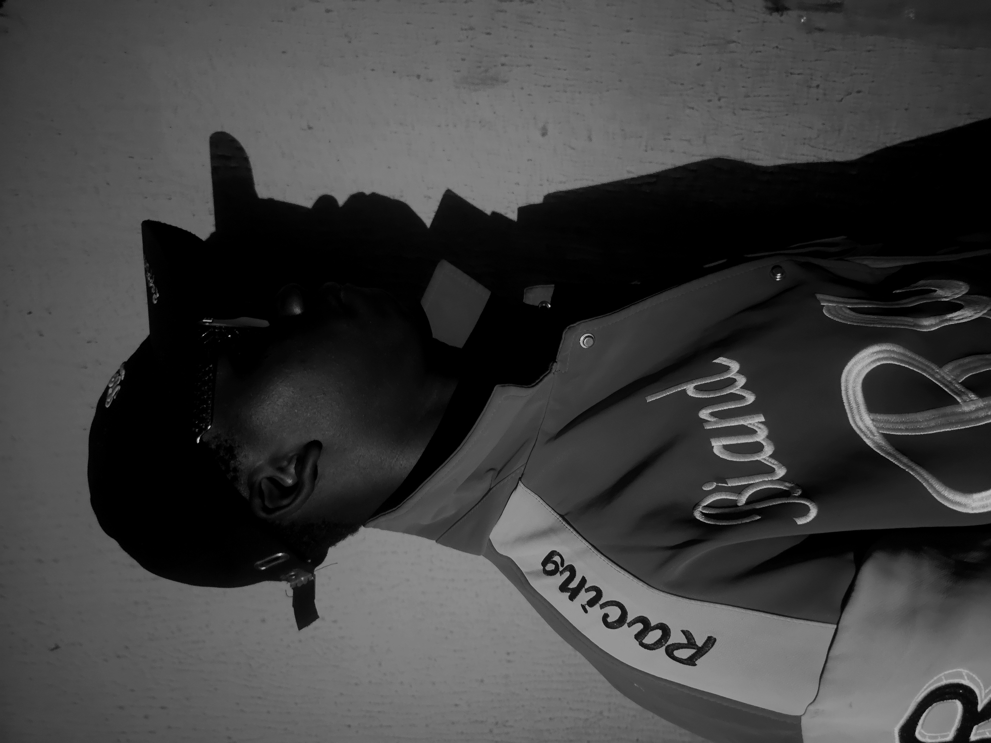A monochromatic, side-angle portrait features a person wearing a dark cap or bandana and a jacket, looking upwards. The individual's face is partially obscured by shadow, with an ear and part of the jawline showing short facial hair. Sunglasses appear to be resting near the ear or on the cap. Strong lighting casts a significant shadow of the person onto a textured, light-colored wall in the background. The shadow includes a distinct shape resembling a raised finger. The jacket is embroidered with text: "Racing" is visible on the sleeve or side, and "Ricard" is prominently displayed on the front in a cursive, stylized font. The scene offers no specific time of day or location cues beyond an indoor setting or a plain background.