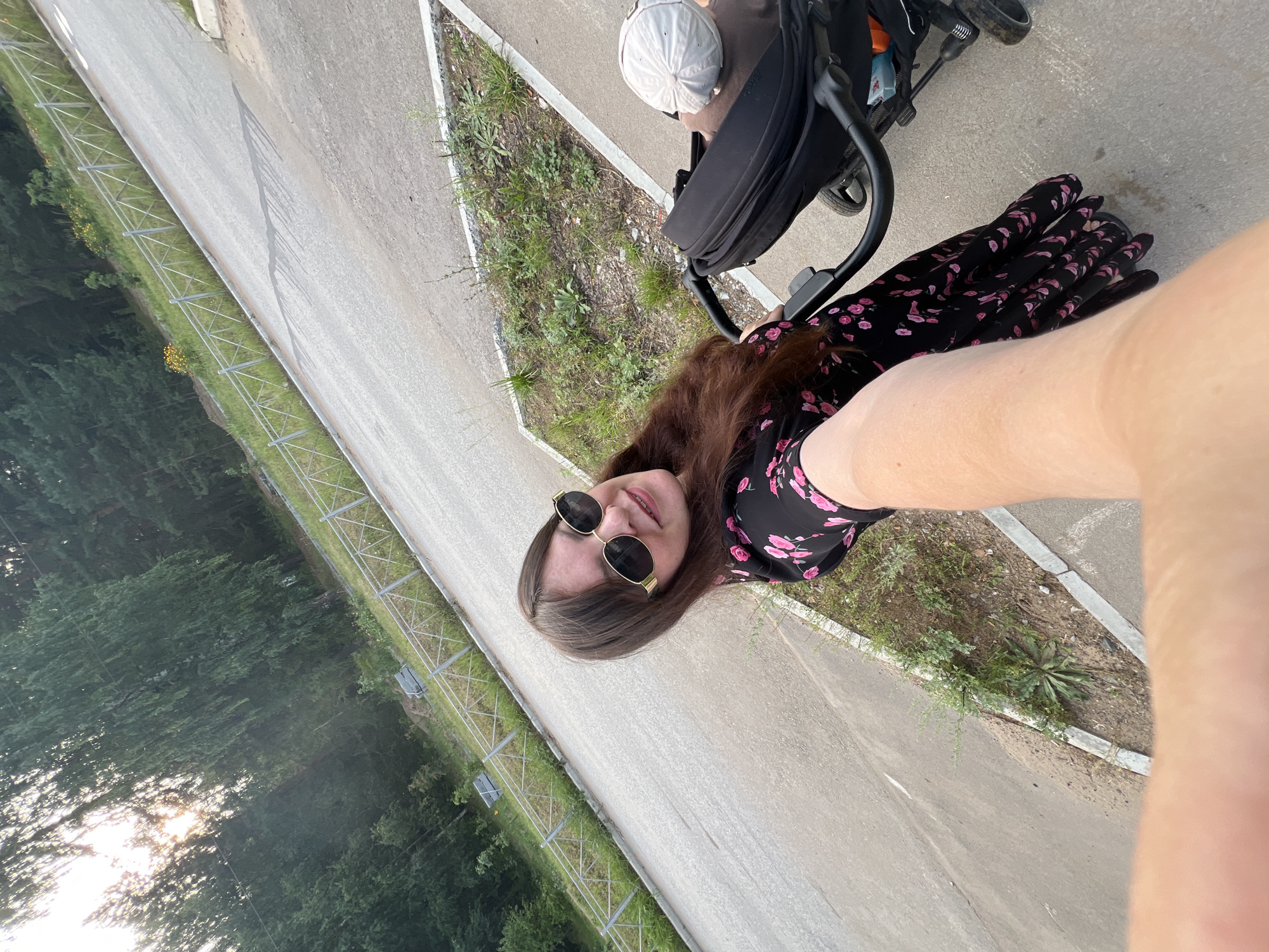 An individual is captured in a selfie taken outdoors, with a stroller visible to their right. The person is wearing a black dress adorned with pink flowers and sunglasses. The background features a paved area with a grassy embankment and a metal fence, suggesting a park or a sidewalk bordering a natural area. Trees are visible in the distance, and sunlight filters through them, indicating it's likely daytime. The scene appears to be a public outdoor space, possibly in Noviy, Russia, given the context provided. The stroller contains a baby wearing a white cap. The perspective of the selfie places the viewer in the position of the person taking the photo, with their arm and hand partially obscuring the lower portion of the frame.