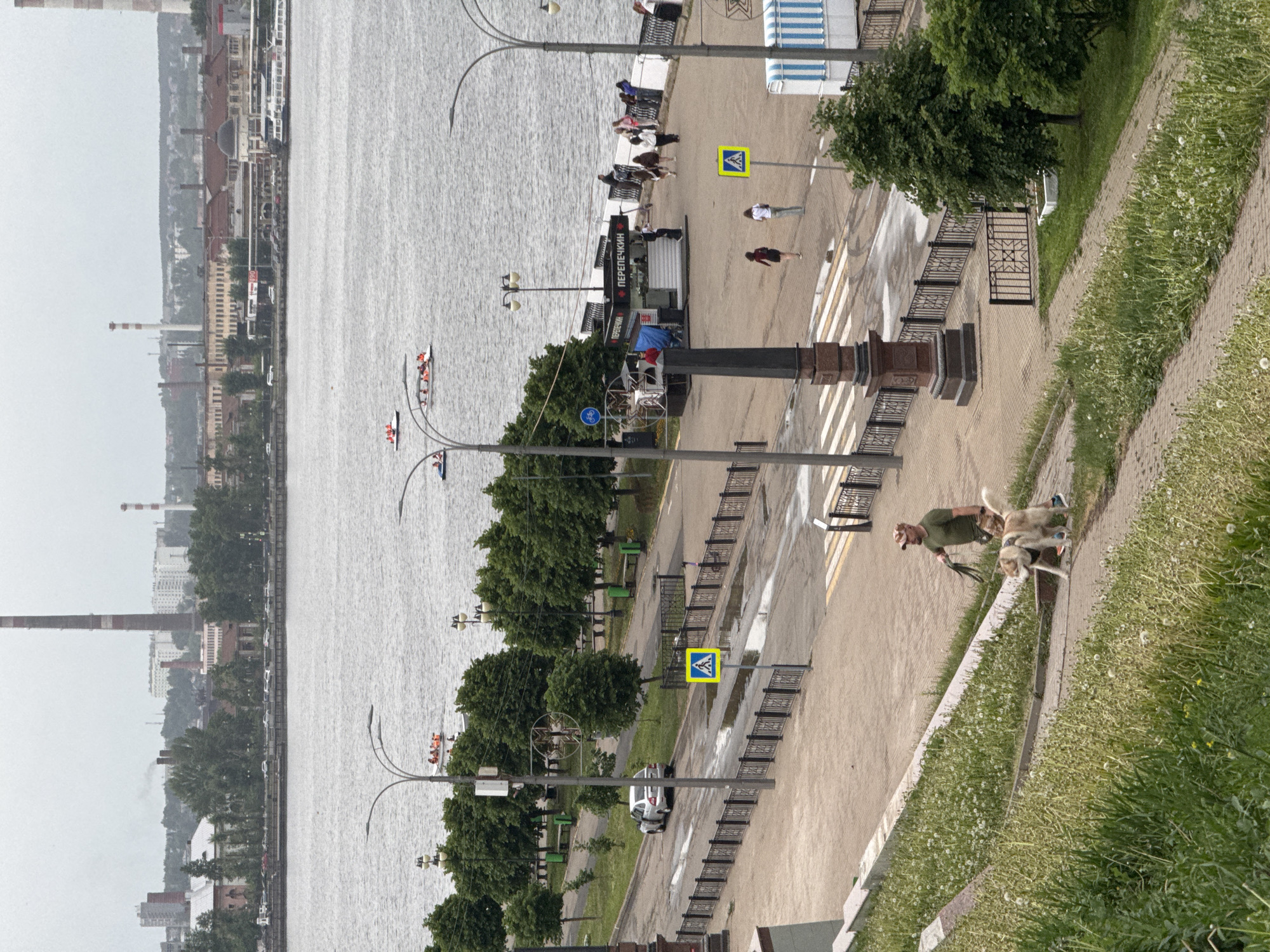 The image shows a scene on the embankment of the Izhevsk Pond in Izhevsk, Russia. The pond occupies the left side of the frame, with buildings and industrial structures visible in the distance along the shoreline. On the right is a paved walkway bordered by a grassy slope. A man walks two dogs along the walkway, near a crosswalk with a pedestrian crossing sign. Further down the walkway, other people stroll. A food stand with the Russian word "ПЕРЕПЕЧКИH" (Perepechkin) on its awning is visible near a crosswalk, where people stand. Several lamp posts are spaced along the walkway. In the background, rowboats are on the pond.