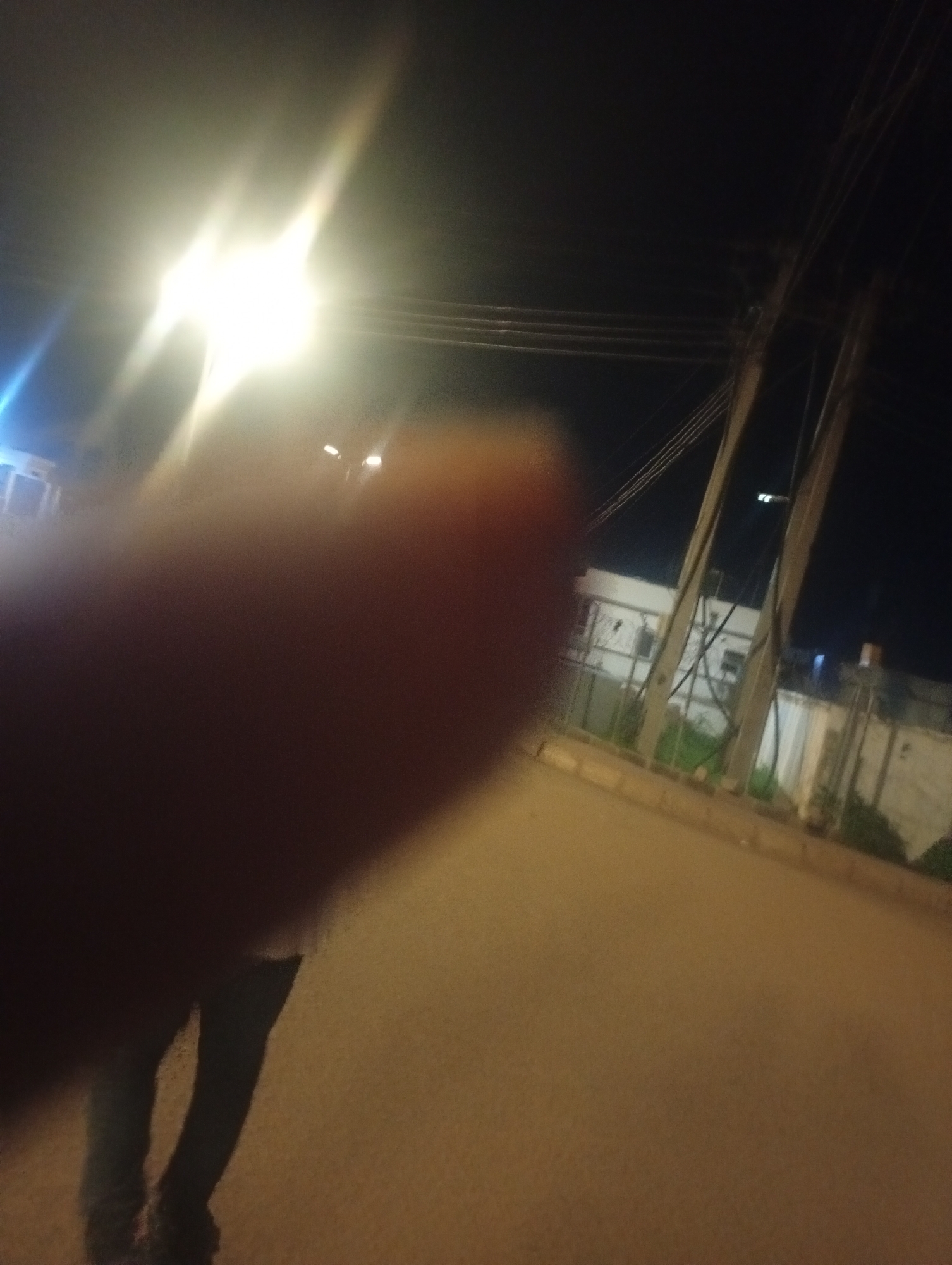 The image depicts a street scene at night in Bukuru, Nigeria. A bright light source, possibly a street lamp or vehicle headlights, is prominently positioned in the upper left quadrant, causing significant lens flare and obscuring details in that area. In the foreground, a blurred finger or hand is visible, partially covering the lower left portion of the frame.
Further into the scene, a person's lower body is visible, appearing to be standing on a dirt or asphalt road. The person is wearing dark trousers and shoes. The road surface is uneven and appears to be made of brown soil or gravel.
In the background, buildings with white walls and windows are visible, along with a fence. Several utility poles with wires are also present, running across the frame. The overall environment suggests an urban or semi-urban setting at night, with artificial lighting creating a dramatic and somewhat hazy atmosphere. The image quality is compromised by low light conditions and the presence of a foreign object (the finger/hand) in the foreground.
