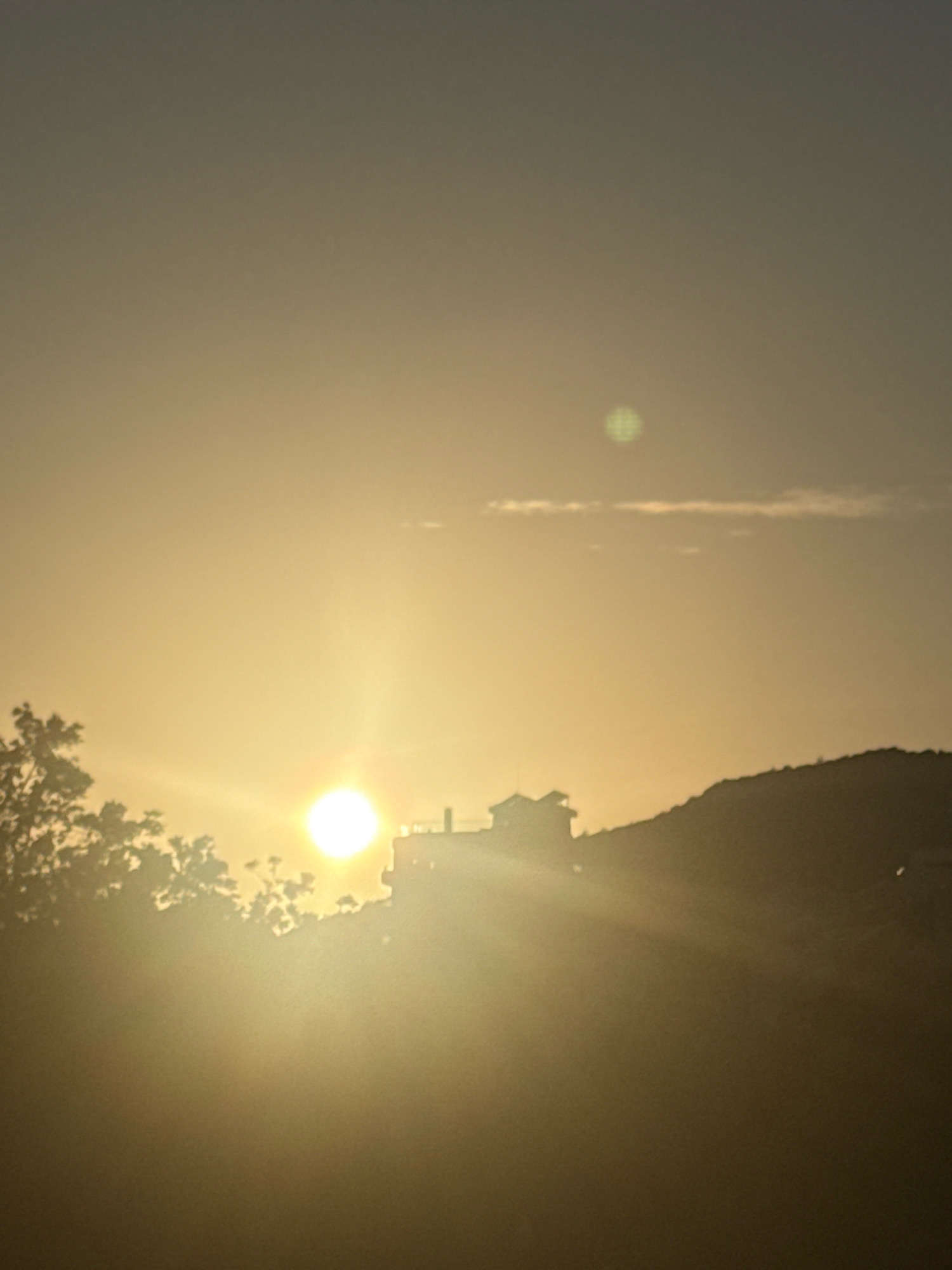 The image captures a serene sunset scene in Boreti, Montenegro. The sun, a bright, glowing orb, is positioned low in the sky, casting a warm, golden light across the landscape. Its rays are visible as bright beams, illuminating the atmospheric haze. In the foreground, the silhouetted shapes of trees on the left suggest lush vegetation. To the right, a hillside rises, silhouetted against the bright sky, with a structure, possibly a house or building, perched on its crest. The sky itself is a gradient of soft yellows and grays, with a faint, hazy cloud formation present. The overall impression is one of tranquility and natural beauty. There are no people or animals visible in the image.