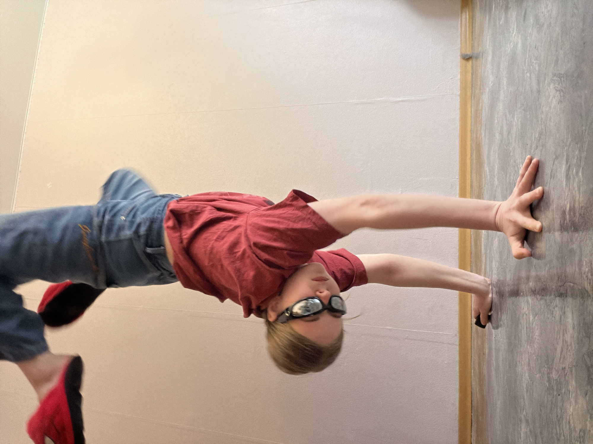 A person wearing jeans, a red t-shirt, red and black footwear, and dark-rimmed glasses is performing a handstand against a wall. The wall is divided into two sections, one painted off-white and the other in a grey marbled pattern. The person is positioned so that their hands are touching the grey marbled section of the wall, while their body is extended towards the off-white section. There is no indication of the location of Perm, Russia.