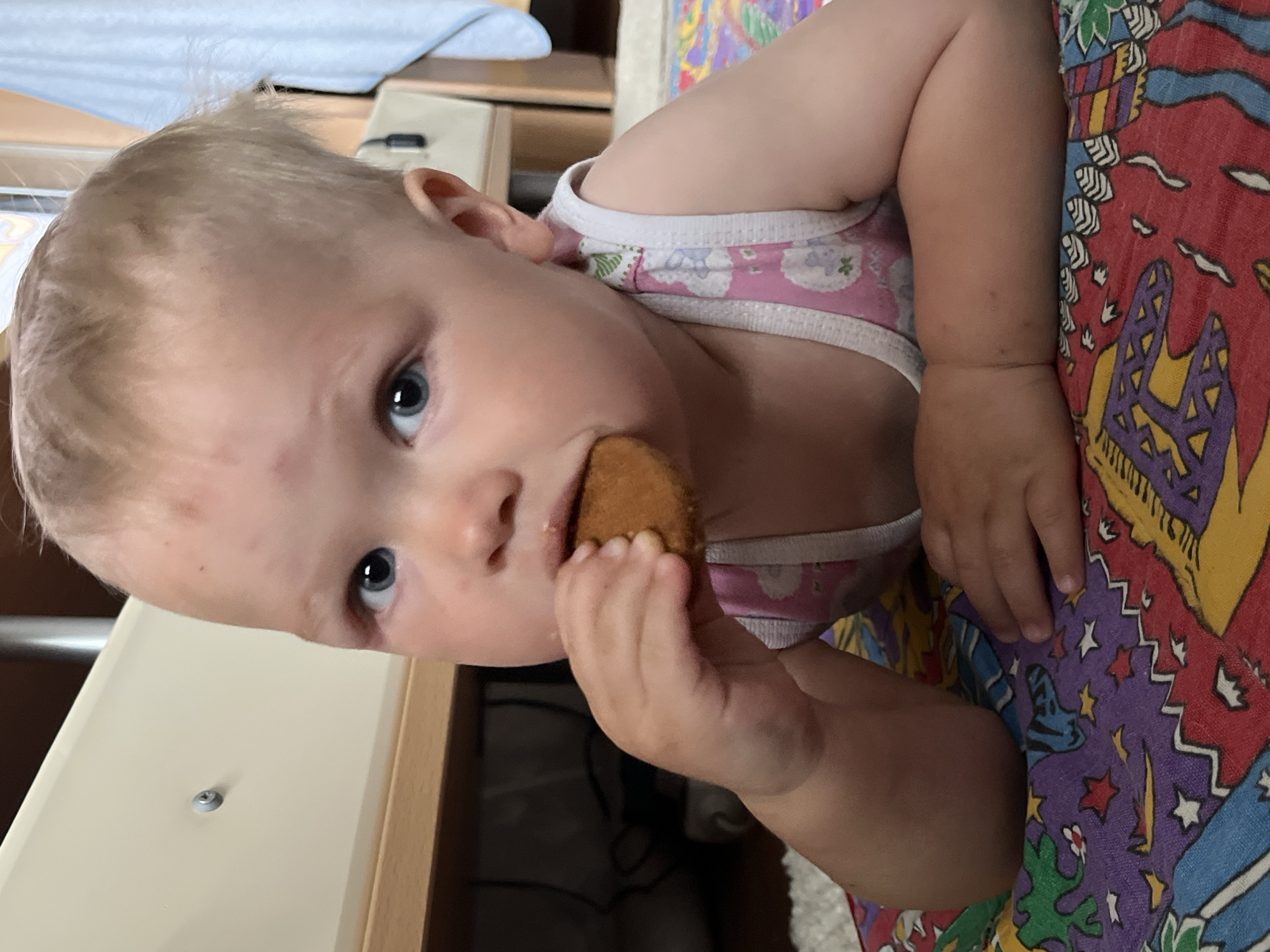 A young child with light blonde hair and blue eyes is eating a round, brown food item. The child is wearing a pink and white tank top. The child's face has some red marks. The background includes a patterned fabric in red, yellow, blue, and purple, a white surface with a metal screw, and a partially visible light blue fabric.
There is no information to determine the location in Chaikovsky, Russia.