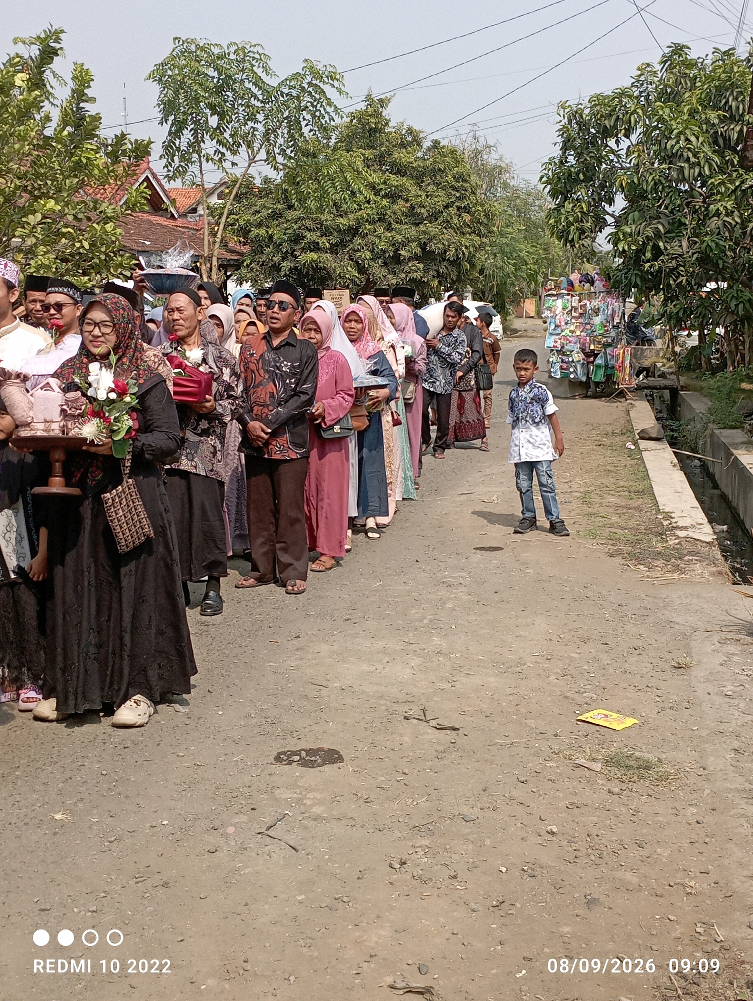 In Dukuhseti, Indonesia, a procession is underway on a dusty, paved road. A group of people, dressed in traditional attire, are walking in a line. The women are wearing headscarves and long dresses, while the men are in shirts and trousers, some with skullcaps. One woman in the foreground is carrying a wooden tray with a floral arrangement and wrapped items. A young boy in a patterned shirt and jeans stands to the side of the road, observing the procession. In the background, trees line the road, and houses with red tile roofs are visible. A vendor's cart with various items is set up on the right side of the road, and a drainage ditch runs alongside it. The sky is clear and bright, suggesting a sunny day. The timestamp on the image indicates it was taken on September 8, 2026, at 09:09.