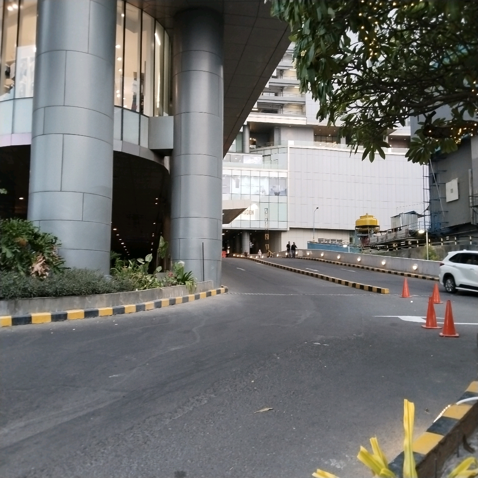 The image depicts an external view of a modern building complex, likely a commercial or residential development, in Surabaya, Indonesia. The foreground features an asphalt road with a black and yellow striped curb. Several orange traffic cones are placed on the right side of the road, and a white SUV is parked partially visible on the far right. In the midground, a curved entranceway leads into a building with large glass windows. The building's facade is characterized by large, cylindrical grey pillars. Beyond this entrance, a multi-story building with a light-colored exterior is visible. Some of the windows in the background are illuminated, suggesting interior lighting. A lush green tree with decorative string lights is visible in the upper right portion of the frame, partially obscuring the background structures. Two figures are walking away from the camera on the right side of the road near the entrance. In the distance, a large yellow water tank is visible on a platform structure. The sky is overcast.