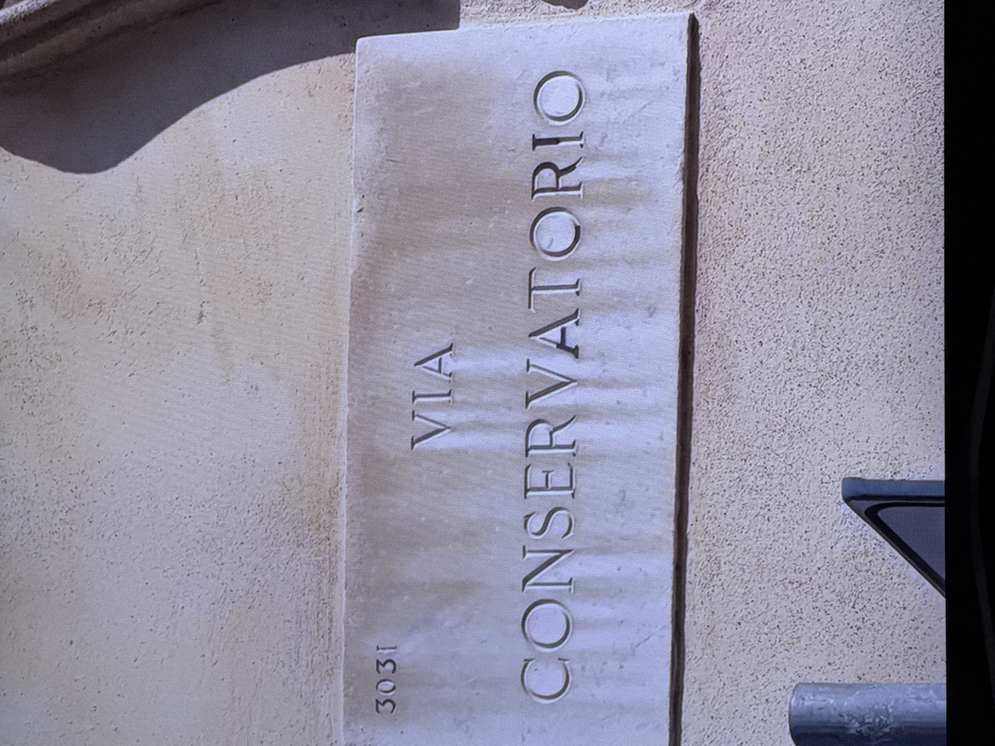 This is an image of a stone plaque mounted on a textured wall. The plaque is rectangular and oriented vertically. It has text carved into its surface. The text reads "VIA CONSERVATORIO" in large, bold, sans-serif capital letters, arranged vertically. Below "VIA CONSERVATORIO" and to the left, the numbers "3031" are also carved into the stone, oriented vertically. The stone appears to be weathered and aged, with some subtle discoloration and texture variations.
The wall behind the plaque is rough and speckled, with a light beige or cream color. To the right of the plaque, a dark shadow and a portion of a dark object are visible, suggesting that the image was taken in a shaded area or with a light source from the left. The lighting in the image is soft, with no harsh shadows, suggesting it might be overcast or taken during softer light hours. There are no people or animals visible in the image. The overall scene is static and architectural, focusing on the informative plaque. The context provided places this scene in Dubai, United Arab Emirates, which is unusual for a street name like "Via Conservatorio" as this is typically an Italian phrase. This suggests the plaque might be part of an art installation, a specific themed area within Dubai, or perhaps a misinterpretation of the location context.