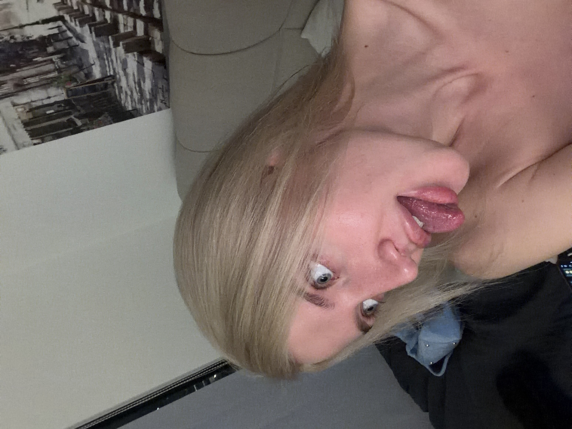 A fair-skinned individual with long blonde hair is depicted in an indoor setting. The subject's head is tilted to the left, and they are shown with an exaggerated facial expression: wide-open eyes directed towards the subject's right and an extended tongue. The upper torso and shoulders are bare. In the background, to the subject's right, a light-colored upholstered headboard or wall panel is visible. Above it, a framed artwork or print depicting a monochromatic street scene with buildings, bollards, and cobblestone-like ground is mounted. To the subject's left, a dark vertical object, possibly a frame, is partially visible. In the lower-left foreground, dark bedding material is present, along with a light-blue cap adorned with metallic studs. The scene is located in Belgrade, Serbia.