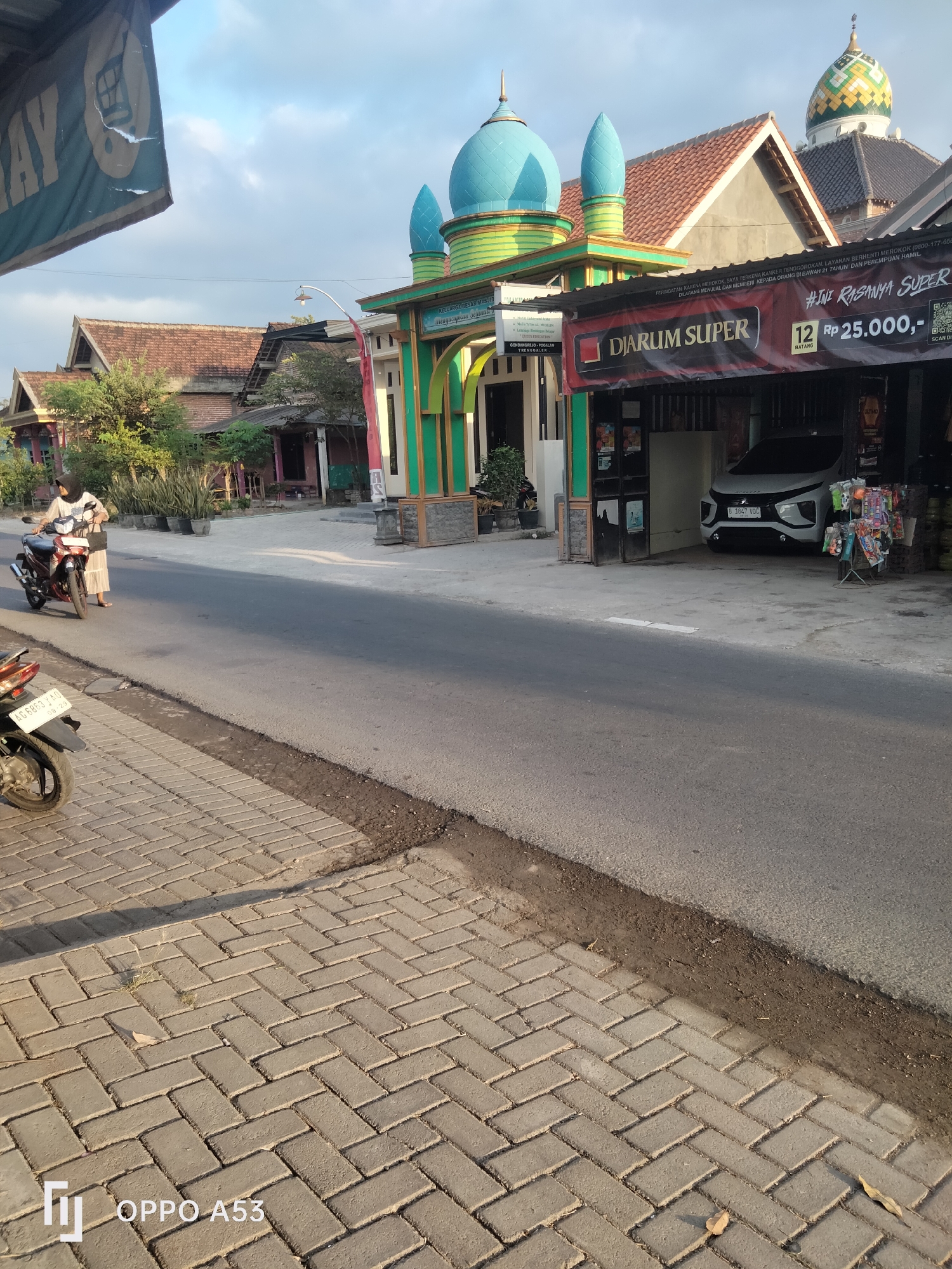 The image depicts a street scene in Trenggalek, Indonesia, bathed in the warm light of what appears to be late afternoon or early morning. On the right side of the frame, a mosque with a striking teal-colored dome and green architectural accents is visible. Adjacent to the mosque, a banner advertising "DJARUM SUPER" is prominently displayed, indicating a tobacco product, and it also states "12 Rp 25.000,-". A white Mitsubishi Xpander car is parked in front of a storefront. A display rack with various small items, possibly snacks or trinkets, is positioned outside the store. On the left side of the road, a woman wearing a hijab and a long dress is standing next to a red motorcycle. She is holding a dark bag over her shoulder. The road itself is asphalt, and a paved sidewalk made of interlocking bricks runs alongside it. The overall environment suggests a typical urban or semi-urban setting in Indonesia, with elements of commerce and religious architecture coexisting.