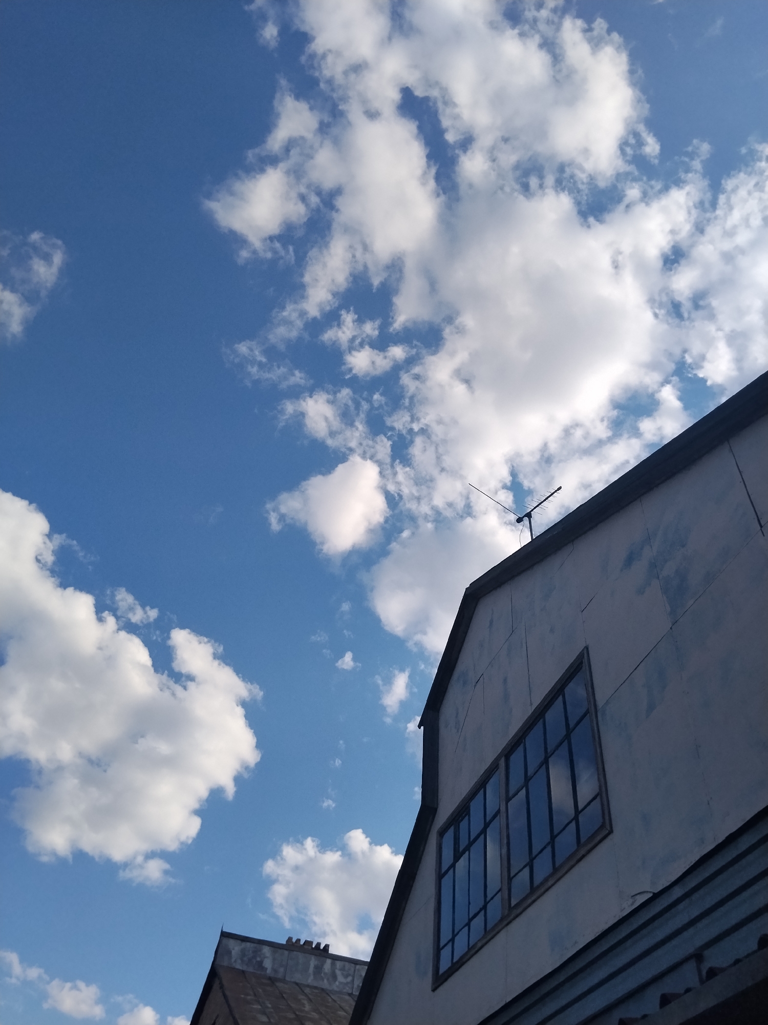 The image shows a view of the sky above a building's roofline. The sky is a clear, bright blue with scattered white, puffy clouds. In the foreground, the upper portion of a building with a sloped roof and a large, multi-paned window is visible. The building's exterior appears to be light-colored and somewhat weathered. On the peak of the roof, a black television antenna is mounted. The lower part of the image reveals another roofline, darker in color, possibly made of corrugated metal, belonging to an adjacent structure. The scene suggests an outdoor view, likely from a higher vantage point looking upwards. The location is identified as Kokshetau, Kazakhstan.