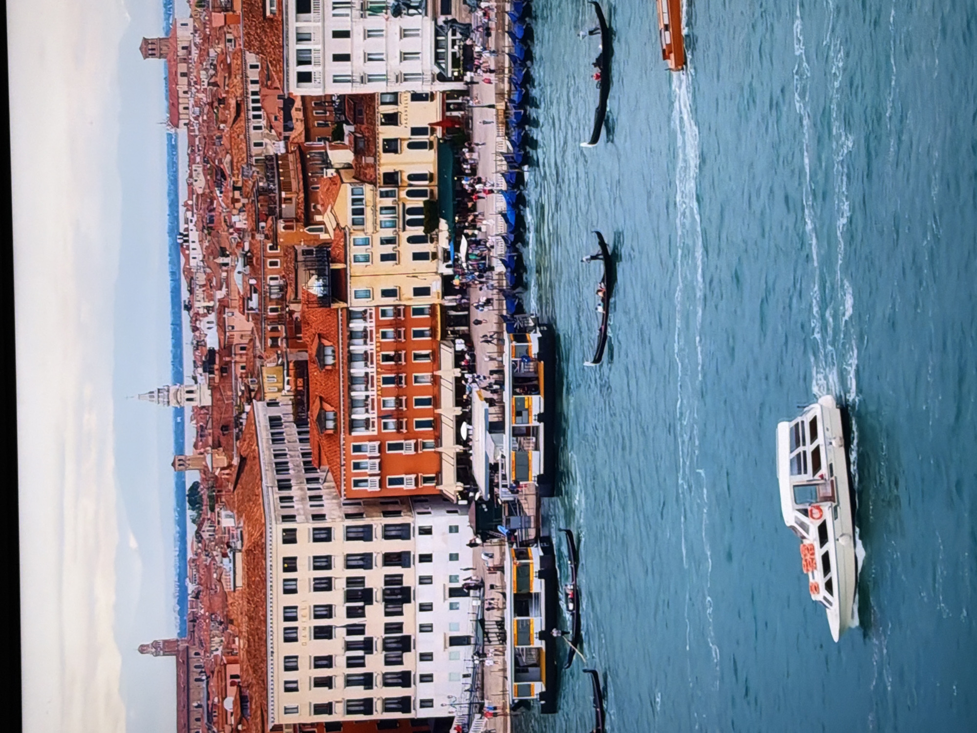 The image depicts a scenic view of a city with canals, boats, and historic architecture. In the foreground, a white passenger boat with a covered cabin is navigating through the blue water, leaving a white wake. To the right, an orange boat is also visible on the water. Several gondolas, each with a gondolier and passengers, are scattered across the canal, some moving and others docked. In the middle ground, two larger passenger ferries are moored along a waterfront lined with buildings. The buildings themselves are multi-story structures with distinct architectural styles, featuring orange-tiled roofs, numerous windows, and decorative facades. A bustling promenade is visible behind the ferries, with many people walking and gathered. In the background, a cityscape unfolds with more buildings and distant mountains under a cloudy sky. The overall impression is of a lively urban environment centered around a waterway. This scene is characteristic of Venice, Italy, not Chirchik, Uzbekistan.
