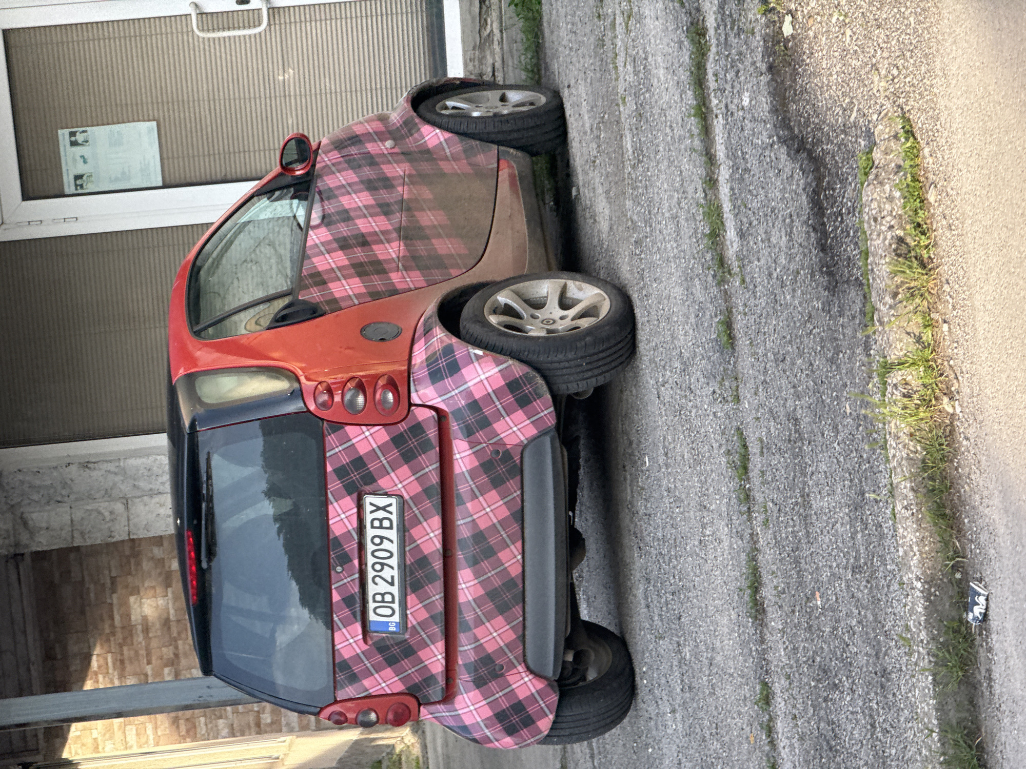 The image shows a red and pink plaid Smart car parked on a concrete surface in Lovech, Bulgaria. The car's license plate number is OB 2909 BX. The surface is cracked and has some grass growing through it. The car is parked close to a building with a closed roller shutter. A notice is visible on the building's facade. The light suggests it is daytime. There are no people visible in the image.