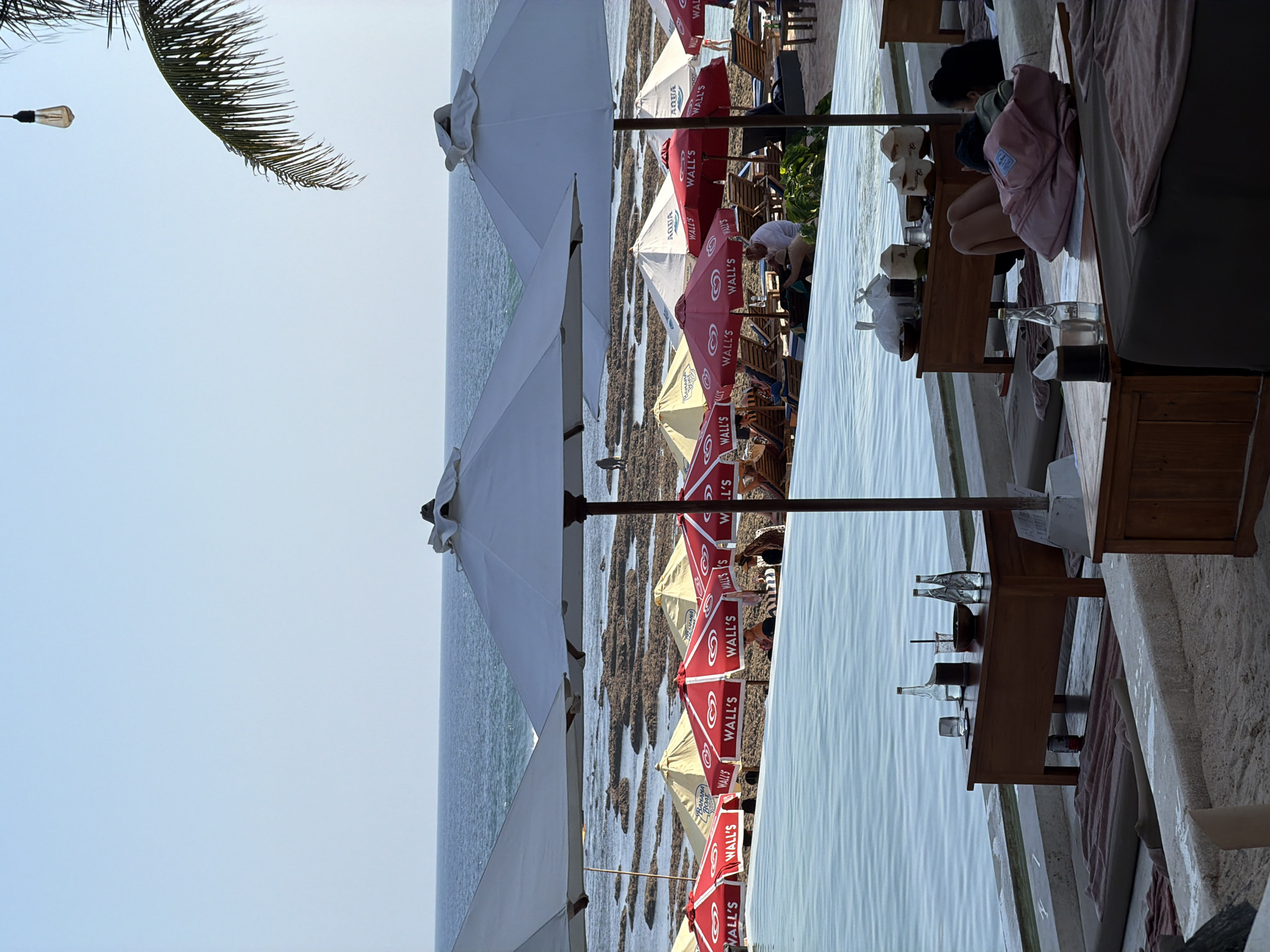 This media file depicts an outdoor seating area at a beachfront establishment, likely a restaurant or cafe, in Kuta Selatan, Indonesia. The scene is set by the ocean, with calm, reflective water occupying the left side of the frame. The foreground and middle ground are filled with rows of beach umbrellas and seating arrangements. Many of the umbrellas are red and white, featuring visible text that includes brand names like "AQUA" and "WALL'S," suggesting a partnership with a beverage or ice cream company. Some umbrellas are a lighter, cream color, and a few large white umbrellas are also present, possibly providing shade for larger groups.
Several people are visible, some seated at tables and others interacting with the environment. One person is seen crouching, possibly attending to something on the ground. Another individual, wearing a pink backpack, is seated at a low table, looking down. The overall atmosphere appears relaxed and leisurely, consistent with a beachside leisure setting. There are wooden tables with various items on them, including bottles, glasses, and what appear to be coconuts, hinting at refreshments being served. The time of day appears to be daytime, possibly late afternoon, given the gentle lighting and the clear sky. The weather seems pleasant and calm, suitable for outdoor dining or relaxation by the sea. The presence of palm fronds in the upper left corner further reinforces the tropical beach location.