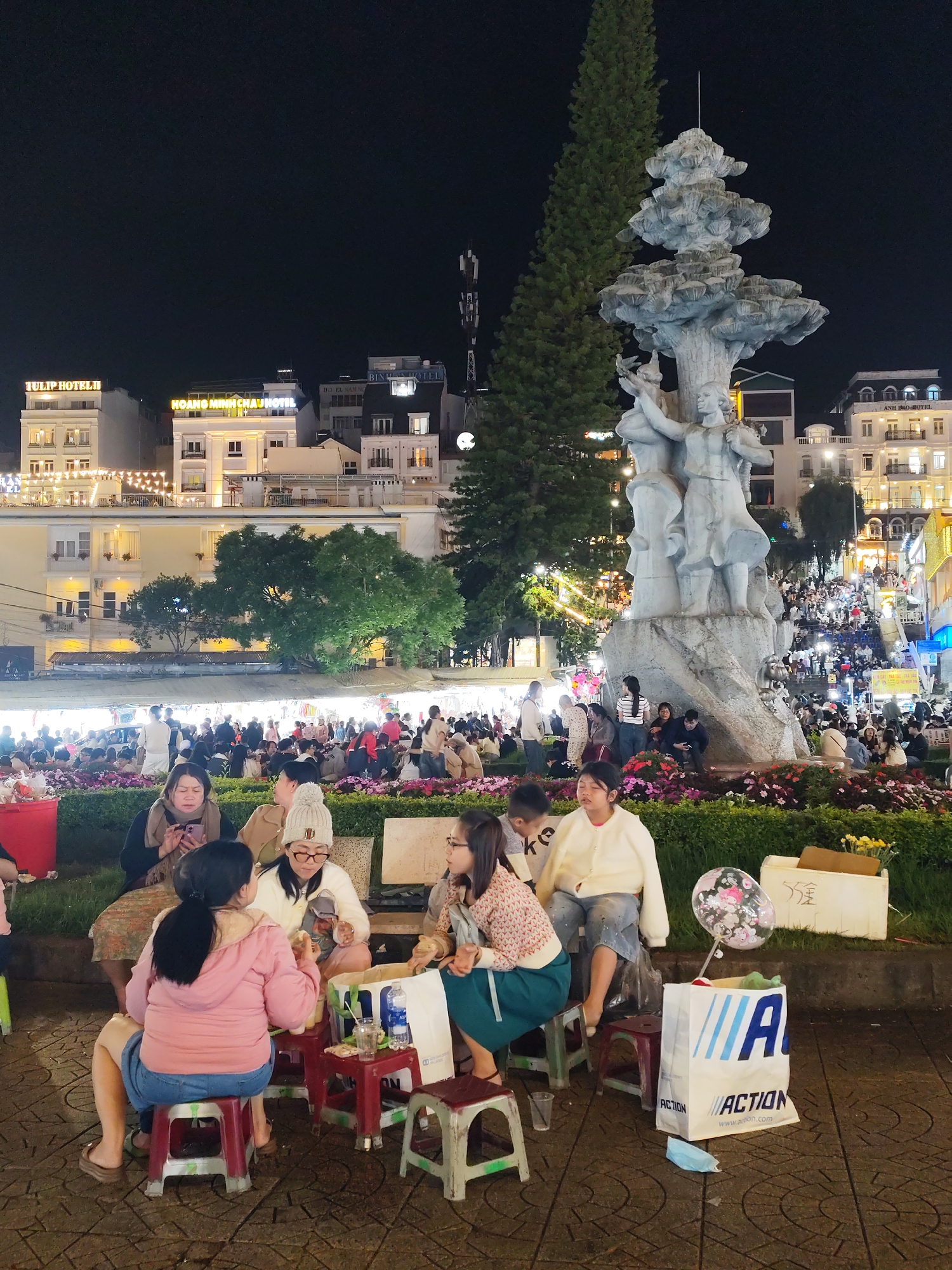 At night in Da Lat, Vietnam, a group of people are seated on small plastic stools around low tables in a public square. They appear to be socializing and eating. The square is crowded with people, and in the background, there are buildings with illuminated signs, including "TULIP HOTEL II", "HOANG MINH CHAU HOTEL", and "ANH DAO HOTEL". To the right stands a tall statue, and next to it is a tall, pointed evergreen tree. Near the group is a shopping bag with "ACTION" printed on it, and a disposable face mask is on the ground. Flowers and other plants are in the vicinity of the statue. The overall scene is bustling with activity, suggesting a lively atmosphere in the city square.
