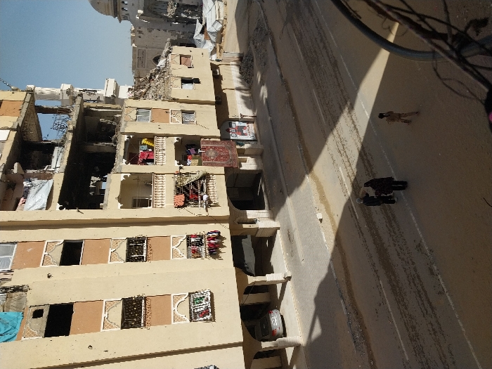 The image depicts a street scene in the city of Khan Yunis, Palestinian Territory, characterized by significant structural damage to buildings. A multi-story, light-colored building dominates the left and center, showing extensive damage from what appears to be shelling or explosions. Multiple floors feature blown-out windows, missing wall sections, exposed interiors, and visible scorch marks. Various items, including clothing and blue tarpaulins, are draped from balconies and windows, indicating continued habitation or attempted repairs. A large red patterned rug hangs from a lower balcony. The building's ground floor features an arched entryway under which a silver sedan is parked.
To the right, another heavily damaged building with rubble is visible, along with a distinct white mosque-like structure featuring a dome in the far background, also appearing affected. Makeshift tents are present in an open, dusty area near these structures. The street below is an asphalt road with visible dirt and debris. Long shadows are cast across the street by the structures. Three individuals are on the street: two adults are standing together, seemingly conversing, while a third, smaller individual walks away to the right. The photograph is taken from an elevated position, with a blurred metal structure in the bottom right foreground. The sky is clear and blue.