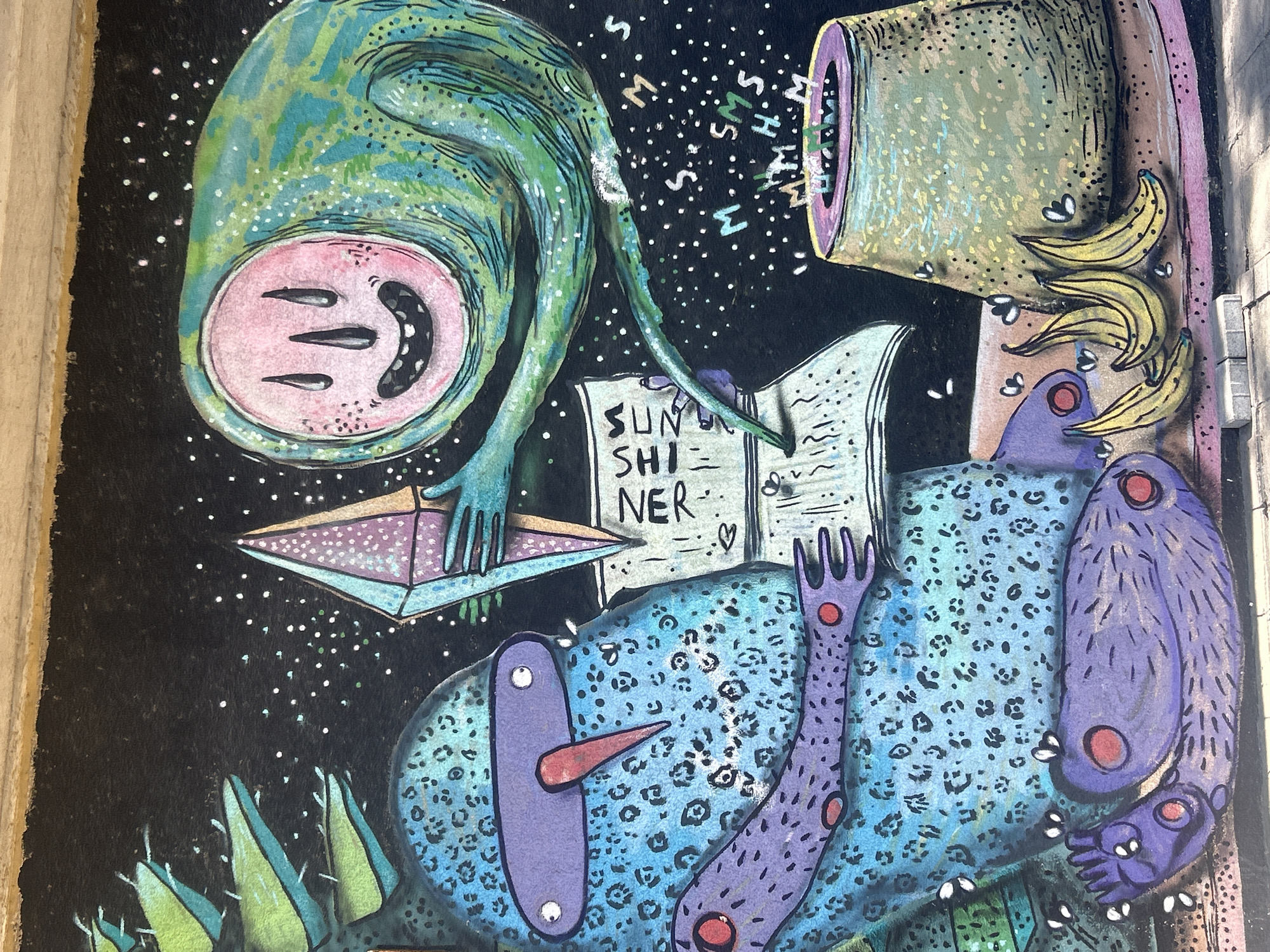 A painted mural on a dark background with light colored polka dots, possibly representing stars. A green and blue upside-down, alien-like creature with a pink, upside-down oval face is on the top left. It has black slits for eyes and a curved line for a mouth with visible teeth. Its long, thin green arm extends to hold a triangular object. The mural contains a cylinder or tube shape, likely ceramic or metal, from which bananas protrude, positioned on the upper right. The mural also includes an abstract creature with blue and purple markings. A book with the word "SUNSHINER" is partially displayed. White letters "M" and "S" are randomly scattered. There is greenery or plant life at the bottom of the mural.