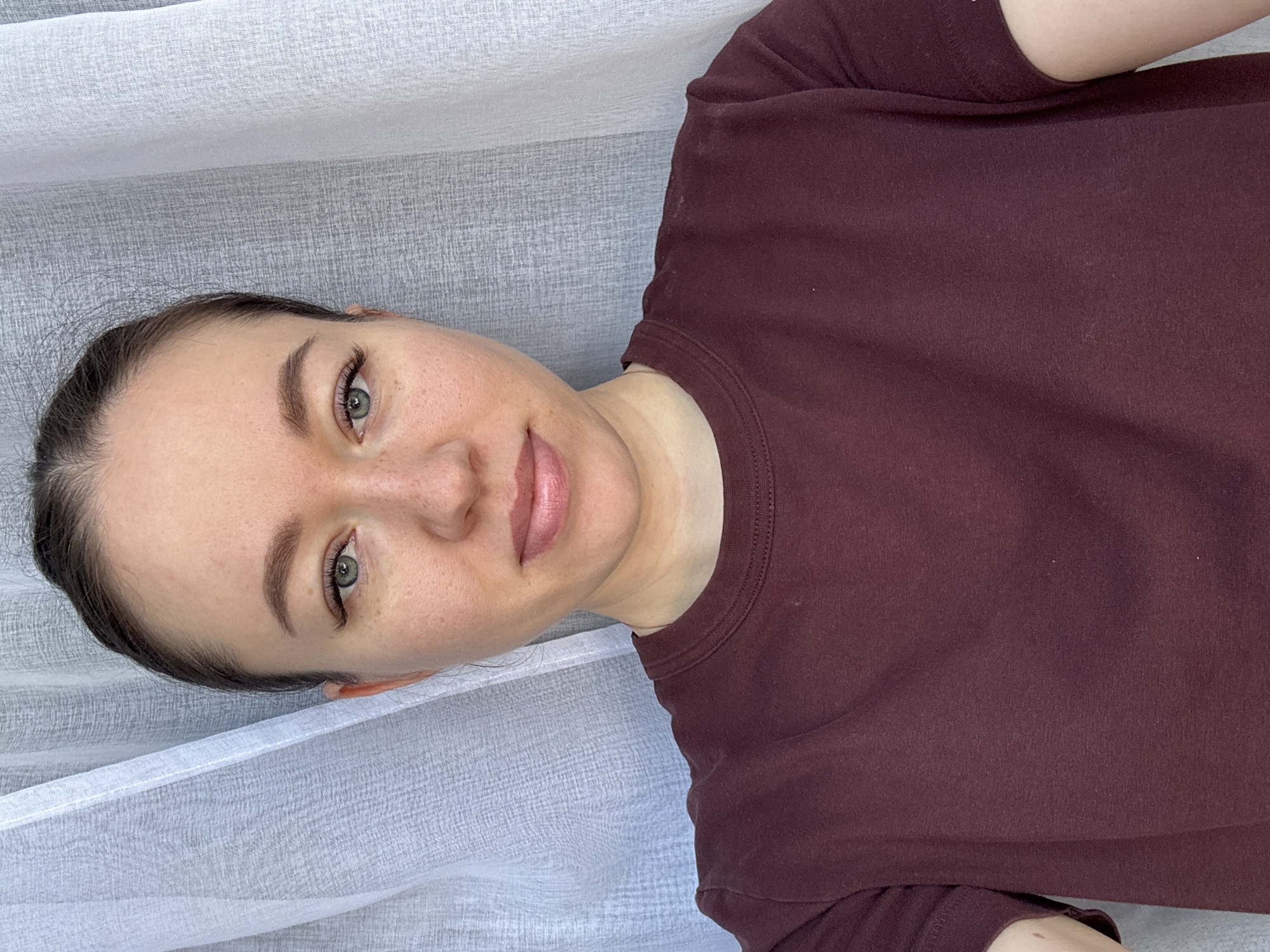 A woman with fair skin, green eyes, and brown hair is lying down, wearing a maroon T-shirt. She is positioned against a textured, light grey or white fabric, possibly a curtain or bedding. The location is not discernible from the image.
