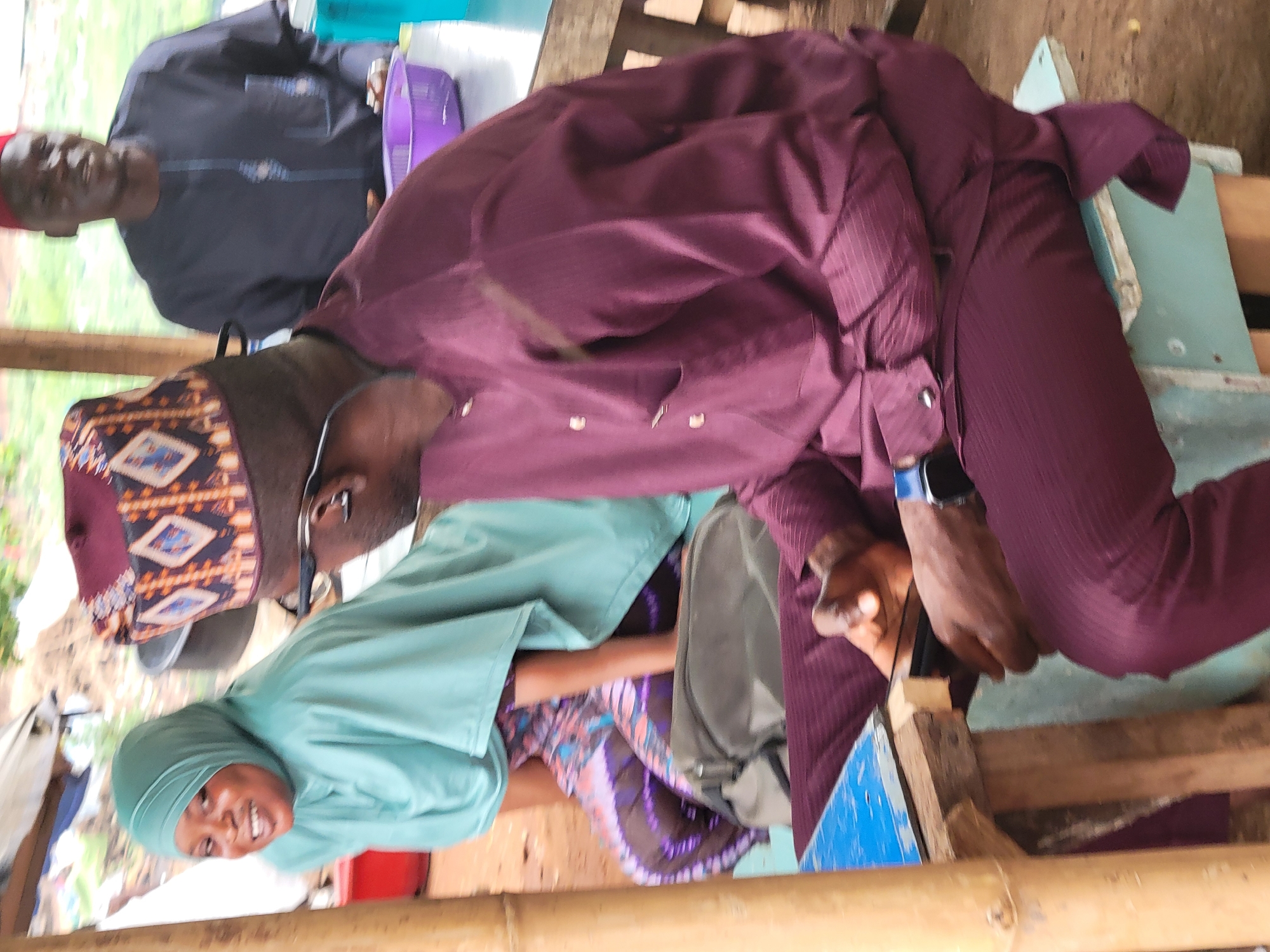 The image depicts four individuals. A woman wearing a light green hijab is smiling at the camera. A man in a patterned hat and glasses is positioned next to her. Another man wearing a dark shirt is slightly behind them to the left. A fourth man in a burgundy outfit is working with his hands on a wooden object. The setting appears to be outdoors with wooden structures and bamboo in the foreground. The location is Abuja, Nigeria.