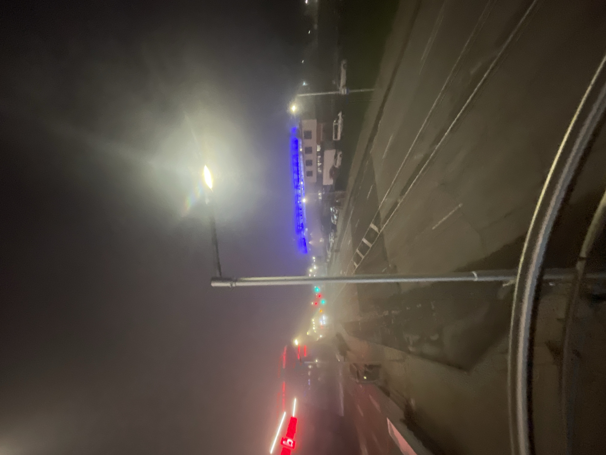 The image shows a view from a high vantage point at night, likely from a window. In the distance, street lights illuminate a fog-obscured roadway. There is a tall building lit with blue lights. There is a light post near the vantage point with a horizontal arm. The red lights of vehicle rear lights and traffic signals can be seen in the haze. The location is likely Burgas, Bulgaria.