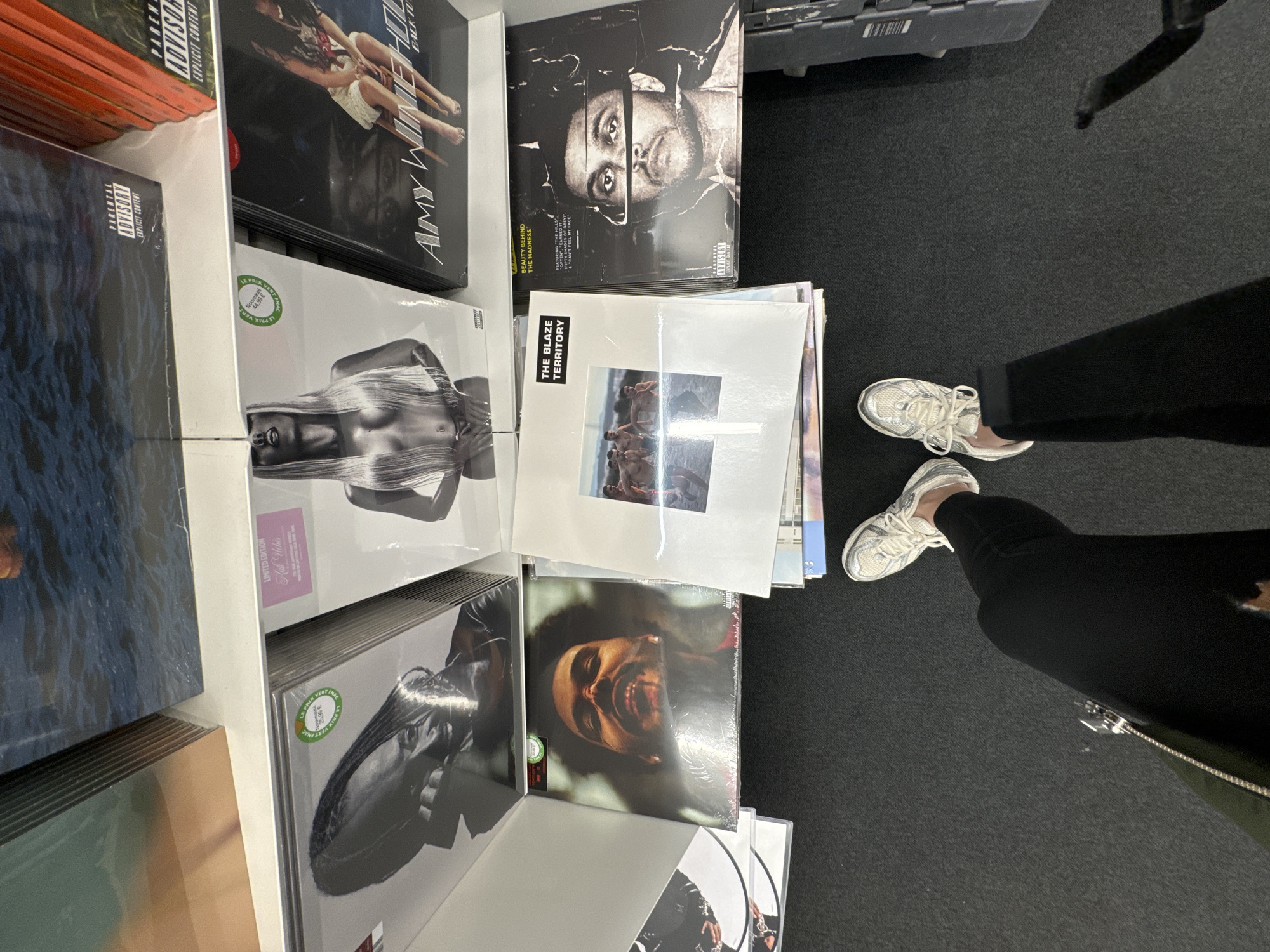 The image shows a person standing in front of a display of vinyl records. The person is wearing black pants and white sneakers. The vinyl records are arranged in rows and feature a variety of album covers, including Amy Winehouse "Back To Black", The Weeknd "Beauty Behind The Madness", and The Blaze "Territory". Some of the records have stickers on them indicating prices in Euros. The environment appears to be a store selling records, possibly located in Paris, France, given the French price tags.