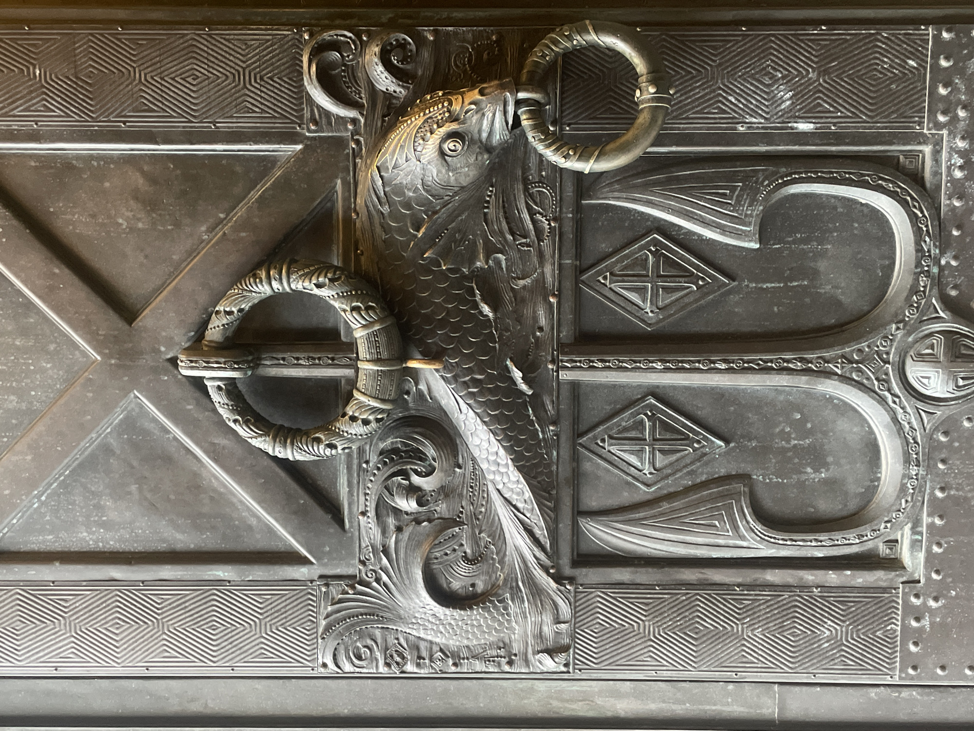 The image shows a close-up of a heavy, ornate metal door, likely made of bronze or iron. The door is divided into several panels with intricate bas-relief carvings. The most prominent feature is a large, stylized fish that spans across the upper central panels. The fish has detailed scales, fins, and a fierce expression, with its mouth open to grip a ring-shaped handle. Above the fish, another ring handle is attached to the door, suspended by a decorative metal band. The panels on either side of the fish are decorated with geometric patterns and scrollwork. The lower panels feature stylized shield-like shapes with diamond-shaped emblems containing cross-like designs. The entire surface of the door is textured with small rivets and weathered marks, indicating age and exposure. This door is located in the Naval Cathedral of Saint Nicholas in Kronstadt, Russia.