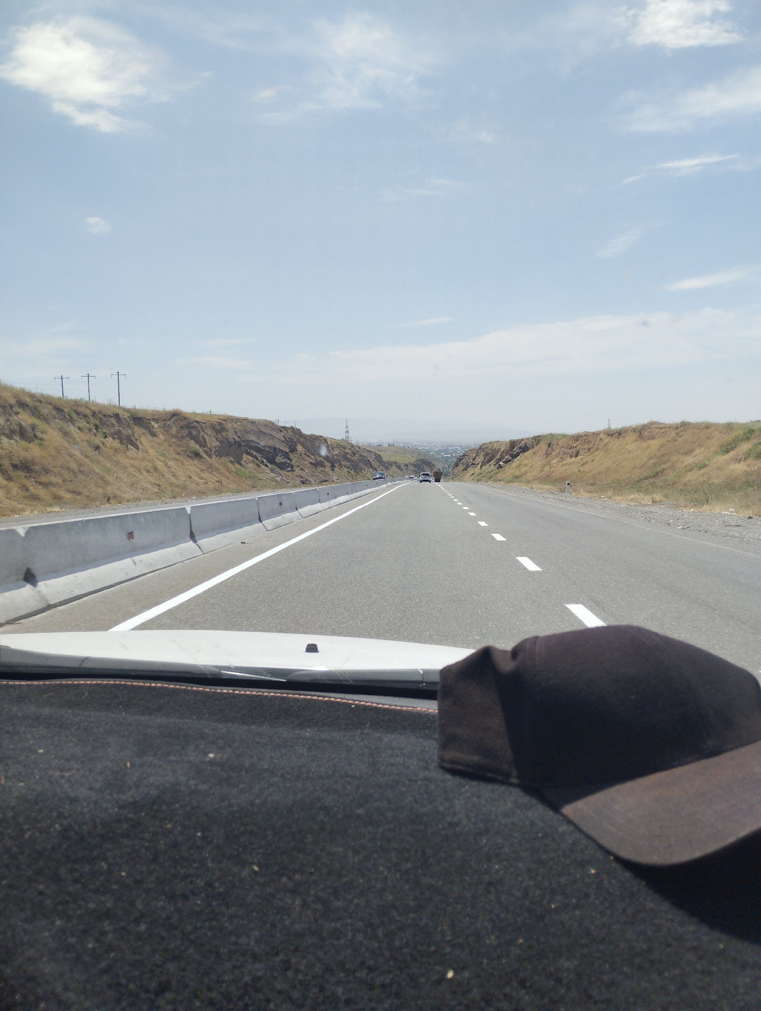 The image shows a view from the interior of a vehicle looking out at a highway. In the foreground, a black baseball cap rests on the dashboard. The highway itself is paved, with white lane markings. To the left of the highway, there is a concrete barrier and a dry, grassy embankment with some rocky outcrops. In the distance, a few cars are visible on the highway, and beyond that, a hazy cityscape can be seen. The sky is light blue with scattered white clouds. The overall environment appears to be dry and sunny, consistent with a warm climate. The location is identified as the city of Sangzar, Uzbekistan.