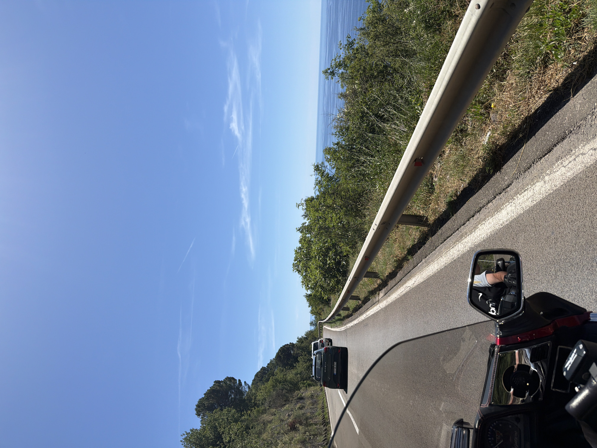 This image depicts a scenic view from the perspective of a motorcycle rider on a sunny day. The road curves to the right, with a steep, tree-covered hillside on the left and a guardrail separating the road from a drop-off to the ocean on the right. The ocean is visible in the distance, a deep blue under a clear sky with a few wispy clouds and contrails. Two cars are driving ahead on the road, with a darker SUV in the foreground followed by a lighter vehicle. The motorcycle's windshield and dashboard are visible in the lower left corner, with a side mirror reflecting the rider wearing a black vest with "bm" logo and black gloves. The environment suggests a coastal highway, likely in a Mediterranean or similar climate, consistent with the provided location of Gornji Katun, Montenegro, which is known for its coastal roads and natural beauty.