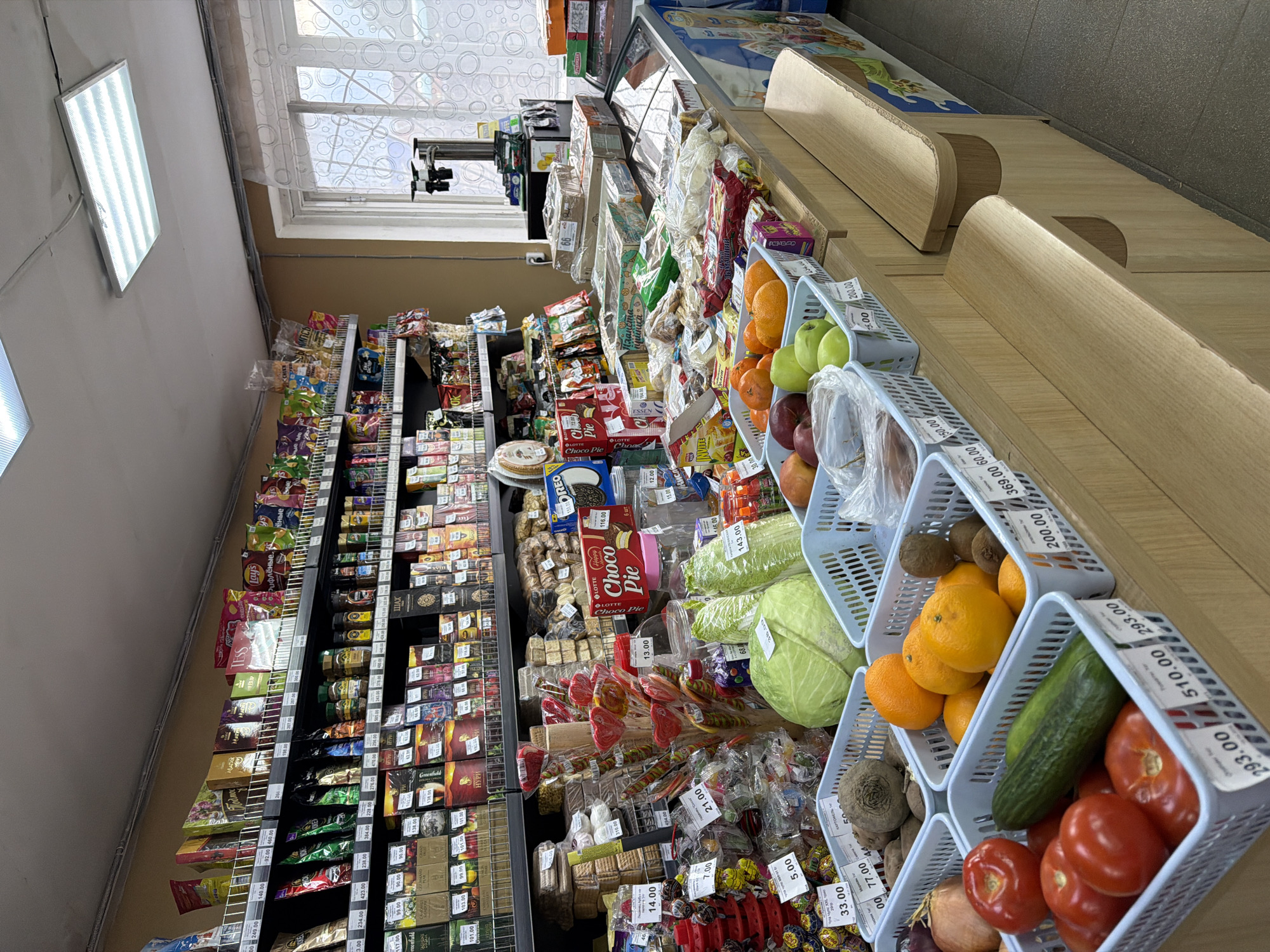 This is an image of a grocery store shelf. It includes shelving units of bagged and boxed food items, and a tiered display of fresh produce such as tomatoes, cucumbers, beets, onions, cabbage, oranges, and apples in light-blue plastic baskets. Price tags are visible near each basket. The store is located in the city of Chaikovsky, Russia. The lighting appears to be provided by fluorescent fixtures in the ceiling and daylight through the window.