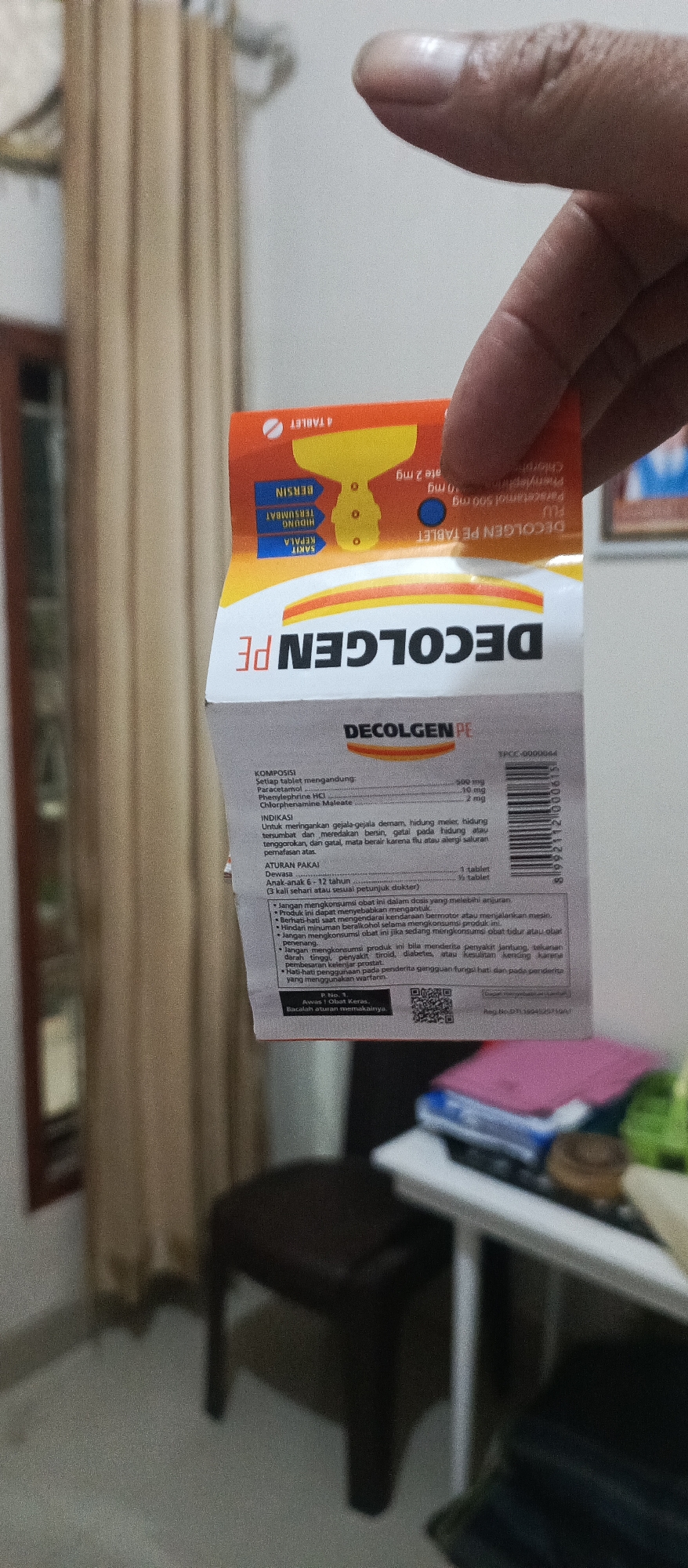 In Arjasa, Indonesia, a close-up shot shows a person holding a small orange and yellow package, likely medication, against a blurred background. The package is partially obscured by the person's fingers, but text such as "DECOLGEN PE" and "4 TABLET" is visible. The package also features graphics of a head, nose, and chest, suggesting it's for cold and flu symptoms. The environment appears to be indoors, with a curtain and a portion of a table visible. The lighting is somewhat dim, and the focus is on the product packaging.
