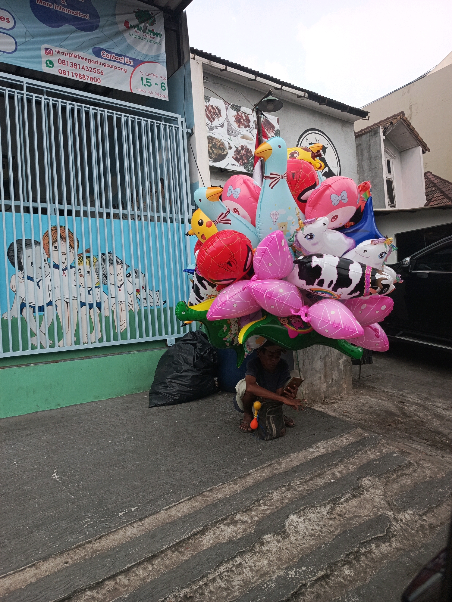 In Pakulonan Barat, Indonesia, a street scene unfolds with a young person squatting on the ground, engrossed in a mobile phone. A large bunch of colorful balloons, including animal-shaped ones and character balloons, are clustered together, seemingly for sale. To the left, a blue metal fence with vertical bars is adorned with a mural depicting children in white and blue outfits, some with pets. A black trash bag rests on the pavement near the fence. A dark-colored vehicle is partially visible on the right side of the frame. The environment appears to be an outdoor setting with a concrete ground and a building in the background.