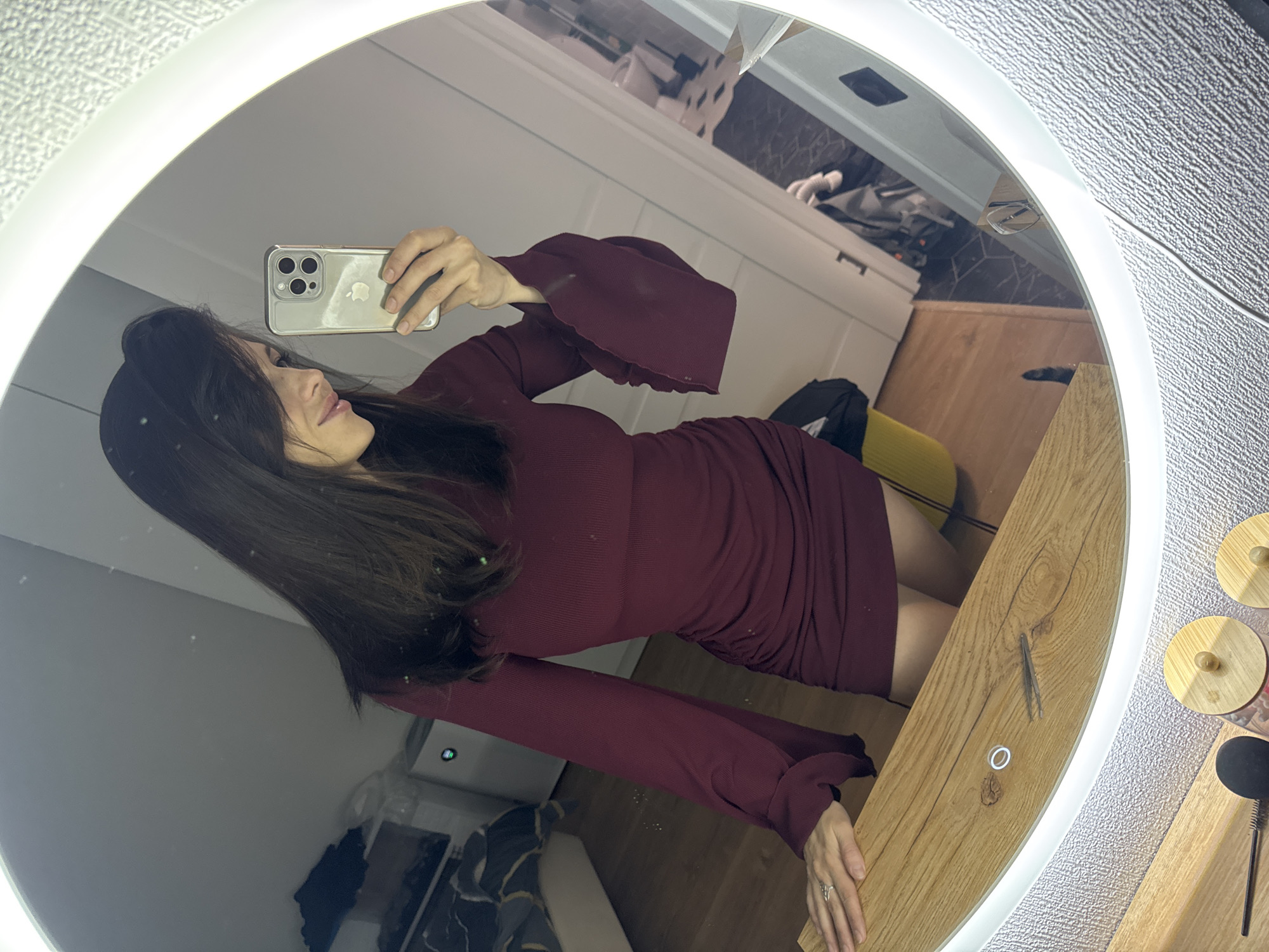 A woman is captured in a mirror selfie, posing indoors in Murino, Russia. She has long, dark hair and is wearing a form-fitting, dark red or burgundy ribbed dress with long, bell-shaped sleeves. A ring is visible on her left ring finger. Her face is seen in profile as she holds up a silver iPhone, which has three camera lenses and a clear case revealing the Apple logo, to take the picture.
The selfie is taken in a round mirror featuring an illuminated white rim, suggesting it's a vanity mirror with built-in lighting. The reflection in the mirror reveals various details of the room. Behind the woman, a white wall is visible. To her right, a light-colored wooden surface or desk holds a white ring and a pair of tweezers. Beyond the mirror's edge to the right, two small, round, wooden decorative objects with knobs are partially visible, alongside a textured, possibly patterned wallpaper.
In the upper reflection, white cabinets with dark handles are seen. Below them, a darker, possibly hexagonal-patterned tiled surface and a light wooden floor are discernible. A yellow object (perhaps a chair or bag) and a dark bag or jacket are also reflected in the background. A white rectangular smart device with a dark display is partially visible behind her left arm. The bright illumination from the mirror suggests an indoor setting with artificial lighting. No other discernible text is visible in the image.