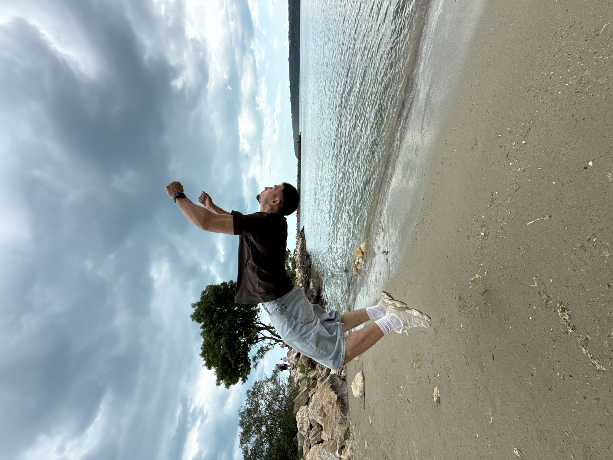 The image shows a man jumping on a beach. He is wearing a dark t-shirt, light-colored shorts, white socks, and sneakers. His arms are raised in the air. The background includes a cloudy sky, a body of water (likely the Black Sea), a sandy beach, rocks, and trees. Given the landscape, this is likely a beach located in Varna, Bulgaria.