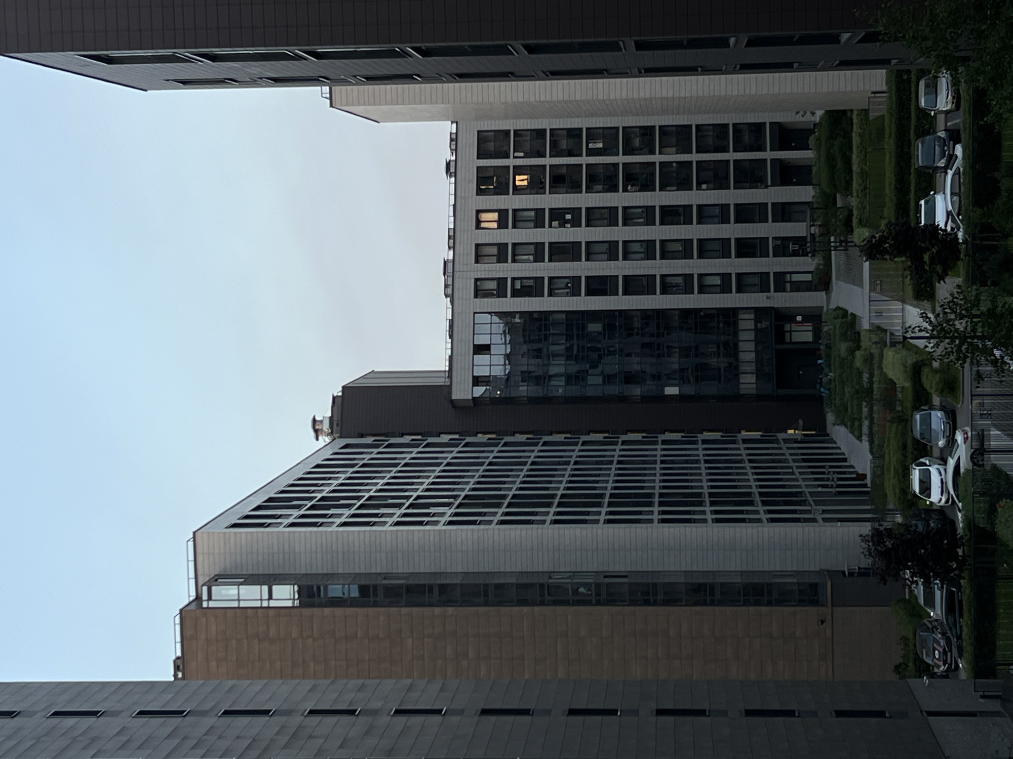 The image depicts a modern urban architectural scene in Saint Petersburg, Russia, characterized by multiple high-rise buildings surrounding a landscaped courtyard and parking area under a light, overcast sky.
From left to right, a partial view of a dark grey building with narrow vertical windows frames the left edge. Adjacent to it stands a taller building with a brown facade on its left section and a light grey facade with a dense grid of square windows on its right. A vertical glass-panel section connects these two material types. Further right and partially obscured, another dark building with reflective glass elements is visible.
The central and right-hand side of the image features a prominent light grey building with a uniform grid of rectangular windows. Several windows on its upper floors show illuminated interiors, with one exhibiting a distinct orange glow. The left section of this building incorporates a large, dark, reflective glass facade. Its flat roof displays multiple small, light-colored ventilation or utility units. The rightmost portion of the image shows another partial dark grey building with vertical window patterns.
At ground level, a meticulously landscaped courtyard is present, featuring manicured green bushes, hedges, and small trees with dark foliage. A paved pathway or drive extends through this area. Below the courtyard, a parking area contains several cars: a black sedan, two dark SUVs, two white sedans, and two silver sedans.
The sky is a pale blue-grey, suggesting either an overcast day or the transition hours of dawn or dusk. The overall lighting is soft and diffuse. No people, discernible actions, or interactions are visible within the frame.
