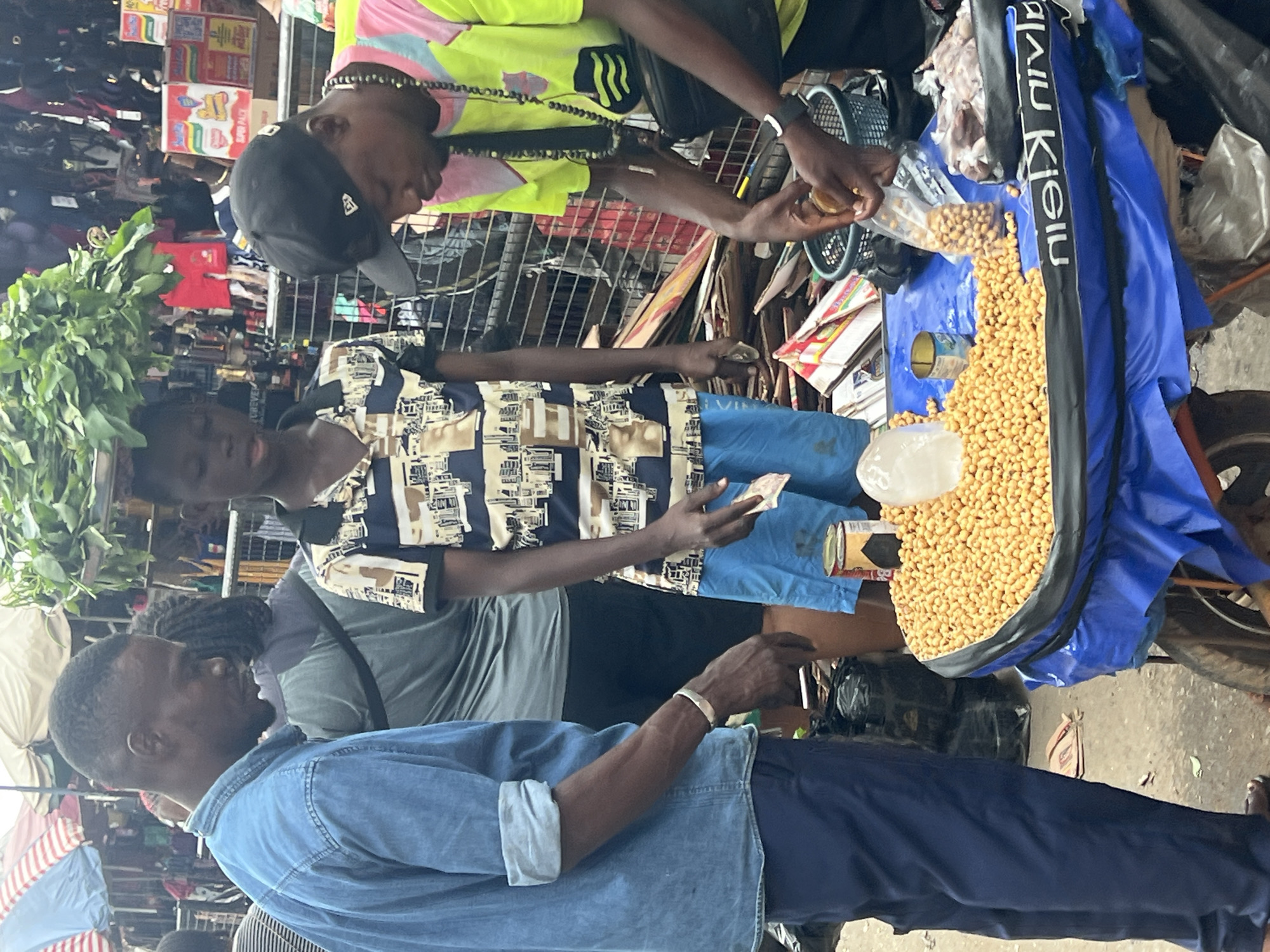 The image depicts a street scene in Nkpor, Nigeria, likely a market. Three individuals are visible. On the left, a man wearing a blue denim shirt and dark trousers stands looking downwards. To his right, a younger individual in a patterned short-sleeved shirt and blue shorts is also looking down, holding a small item in their right hand. On the far right, a person wearing a brightly colored shirt and a black baseball cap is actively engaged, holding a plastic bag filled with small, light-colored seeds or nuts. They are manipulating the bag over a black basket. In the foreground, a table covered with a blue tarp displays a large pile of these same seeds or nuts, along with a clear plastic bag containing more of them, a can, and a block of ice. The background is cluttered with market stalls, merchandise, and other people, suggesting a busy commercial environment. The overall impression is one of daily commerce taking place in an open-air market.