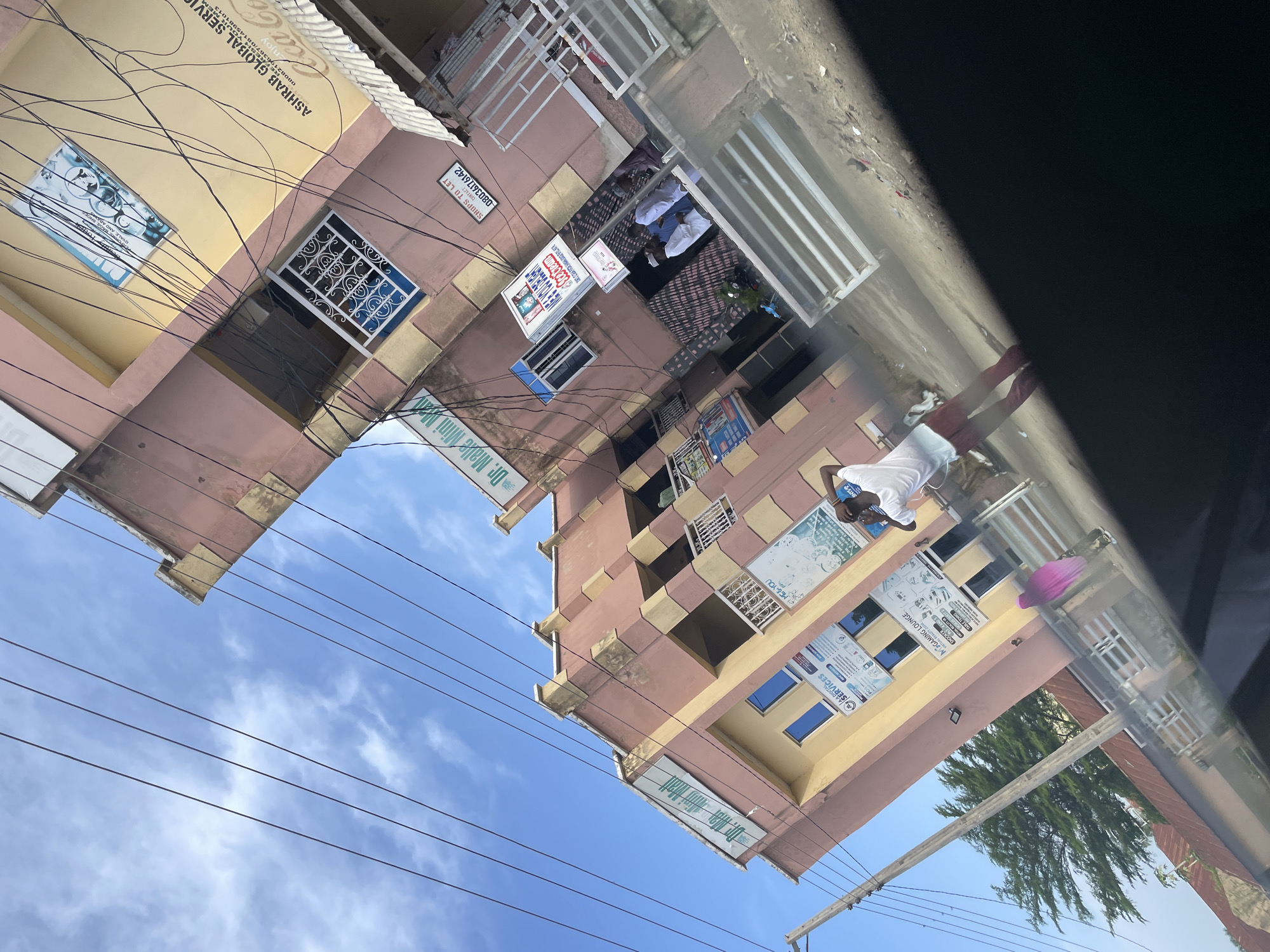 The image depicts a street view in Jalingo, Nigeria, on a bright, cloudy day. Two multi-story buildings, primarily painted in hues of pink and yellow, dominate the scene. The buildings are adorned with various signs, indicating businesses such as "ASHRA GLOBAL SERVICES," "Dr. Malle Mini Mall," and "MC GAMING LOUNGE." Overhead, a tangle of electrical wires crisscrosses the sky, casting shadows on the buildings.
In the foreground, a person in a white t-shirt and red trousers is seen standing with their back to the camera, appearing to stretch or adjust their clothing. Further down the street, another person, wearing a pink headscarf, is visible walking. The ground is a mixture of paved surfaces and dirt, with some debris scattered around. The architectural style of the buildings suggests a blend of modern and functional design. The overall impression is a bustling urban environment, characteristic of many Nigerian cities.