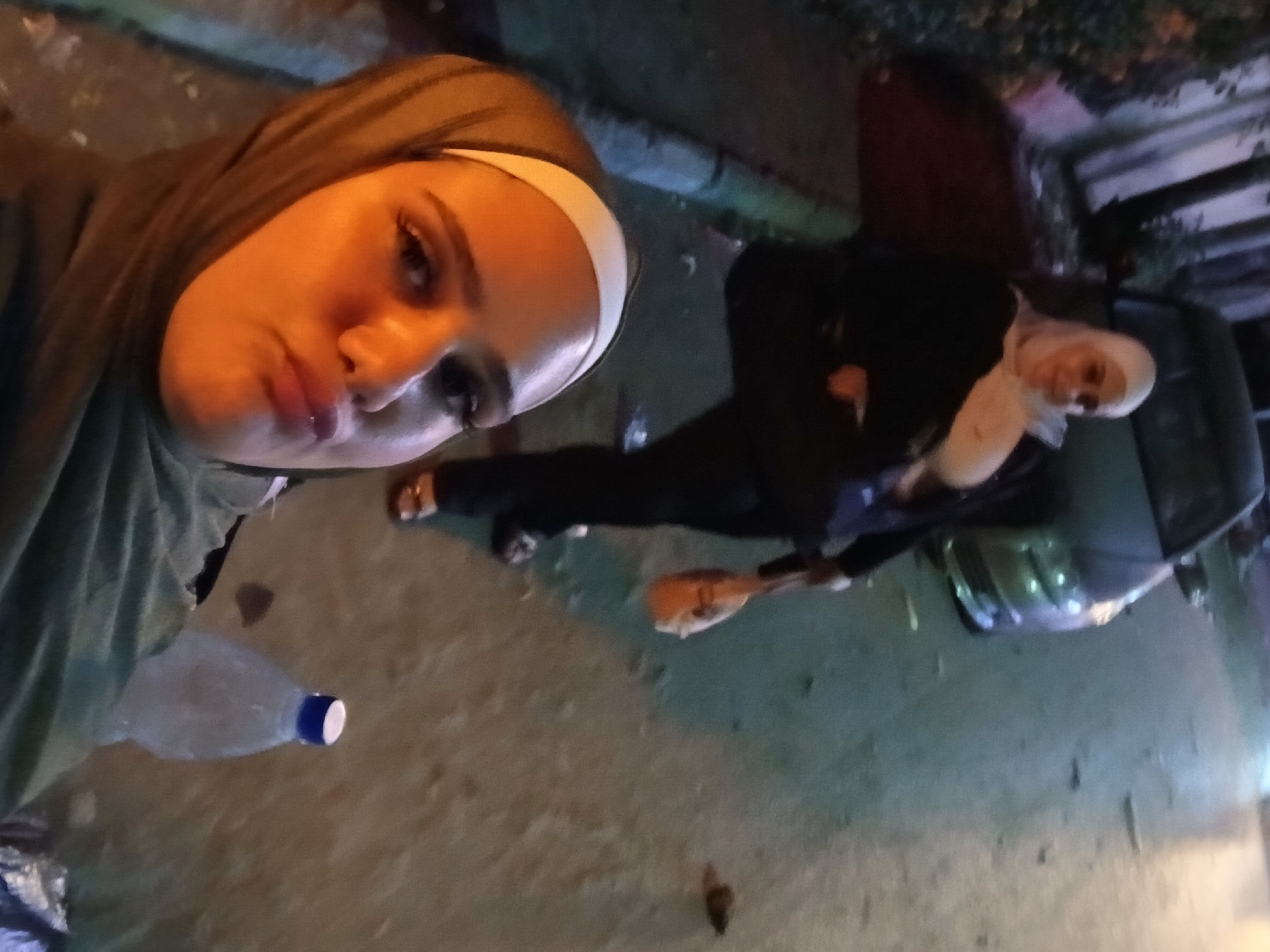 The image captures a candid moment on a street in Qudsaiya, Syria, at night. In the foreground, a woman wearing a green hijab and a white headband makes a pouty face towards the camera. Her eyes are wide open, and she has noticeable eyelashes. Behind her, a second woman, also wearing a white hijab, is partially visible, looking towards the camera with a slight smile. She is dressed in a black jacket and appears to be holding an orange bag. A dark-colored car is parked on the right side of the street. The ground is paved and appears somewhat littered. The lighting suggests artificial streetlights or ambient light sources are present.