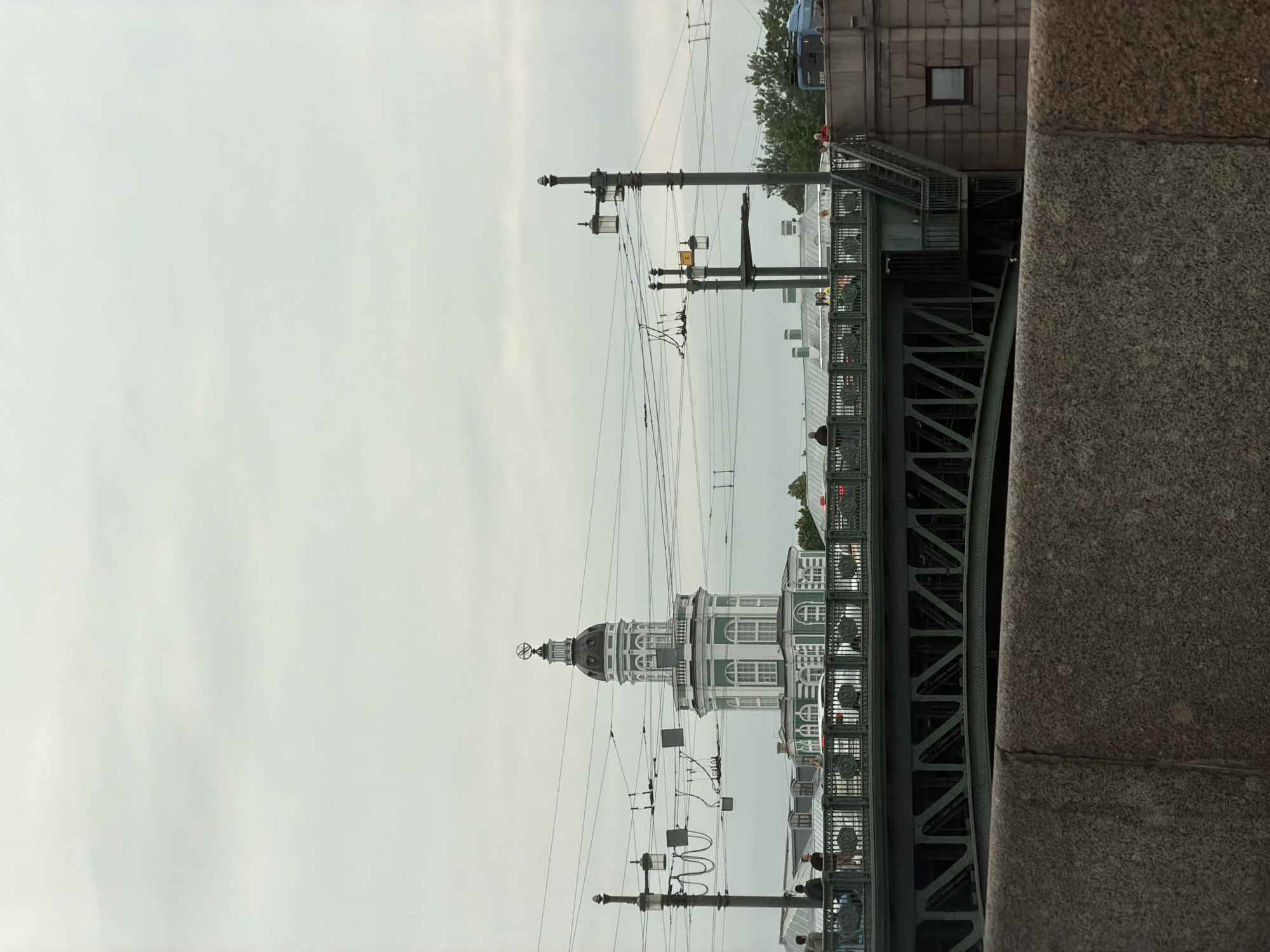This is a medium shot photograph of the Kunstkamera building in St. Petersburg, Russia, on a cloudy day. The Kunstkamera is a distinctive tower-like structure with green and white detailing, topped by a dome and a spire with a globe. The tower is seen from across a bridge, with the bridge itself taking up a significant portion of the foreground and midground. The bridge is a dark metal structure with intricate railings and visible support beams. Several people are visible on the bridge, some standing and looking around, others walking. Overhead, a complex network of electrical wires for trams or trolleybuses stretches across the sky, supported by poles. To the right of the Kunstkamera, more of the bridge structure and a stone building with a small window are visible, along with trees and a blue vehicle, possibly a tram, in the distance. The foreground is dominated by a stone wall, partially obscuring the view of the bridge.
