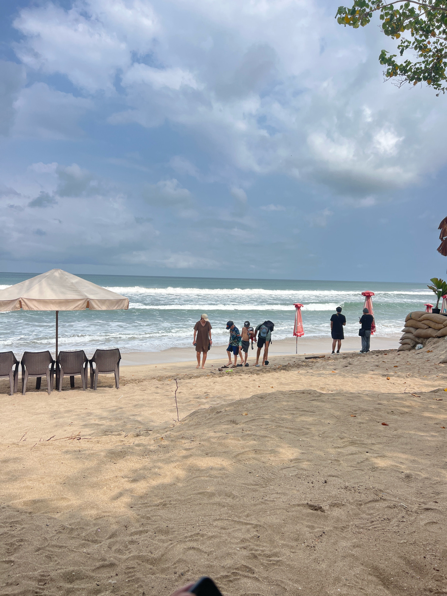 A group of people are on a beach in Kuta, Indonesia. The sky is cloudy, and there are waves in the ocean. In the foreground, there are chairs and an umbrella. In the middle ground, there are several people standing and walking on the sand. In the background, there are more people and some sandbags. The overall scene is one of a beach on a cloudy day.