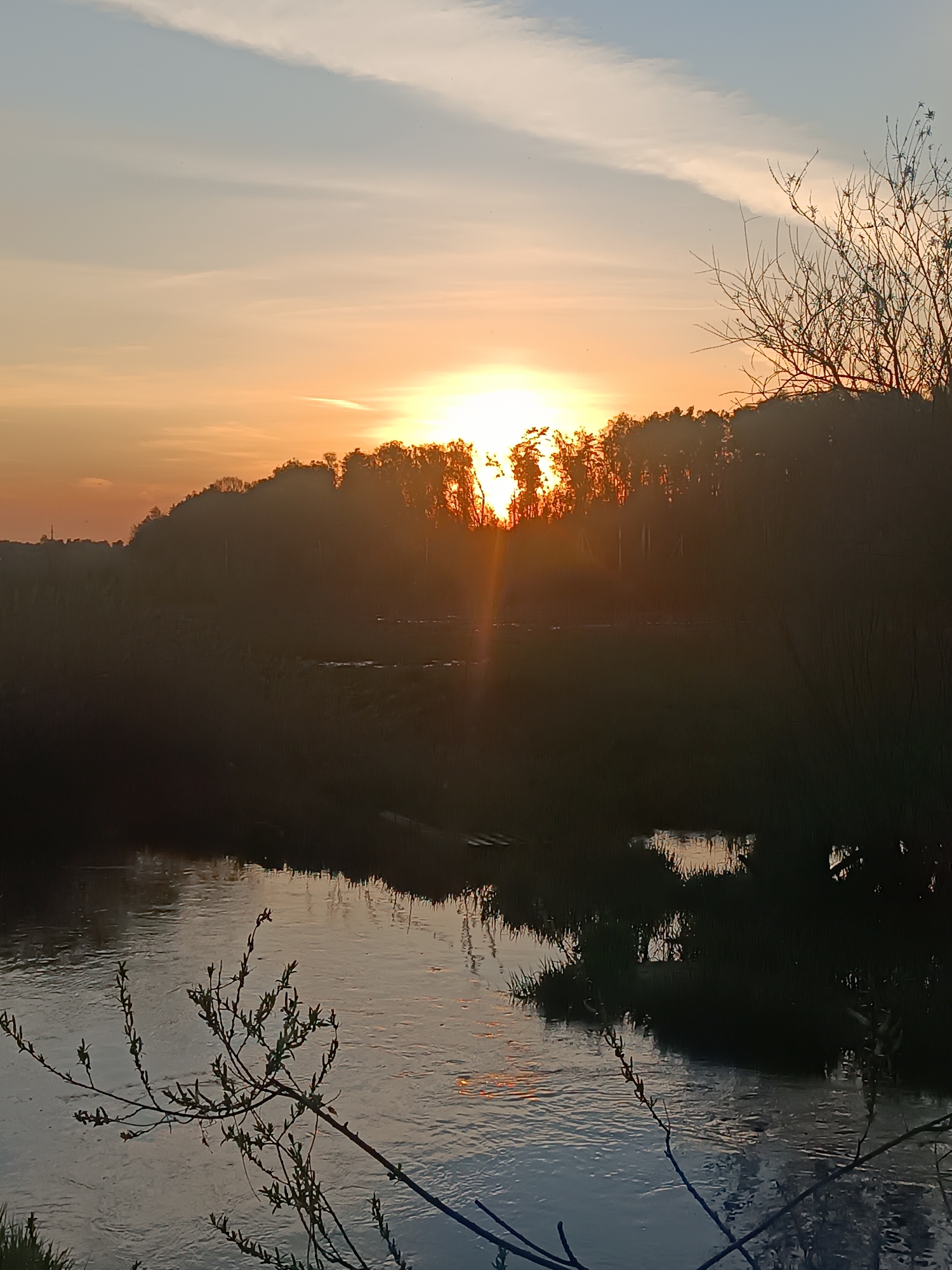 The image depicts a river at sunset. The sun is setting behind a line of trees in the background, casting an orange glow across the sky. The river reflects the colors of the sunset. There are branches with emerging leaves in the foreground. The location is Unknown, Unknown.