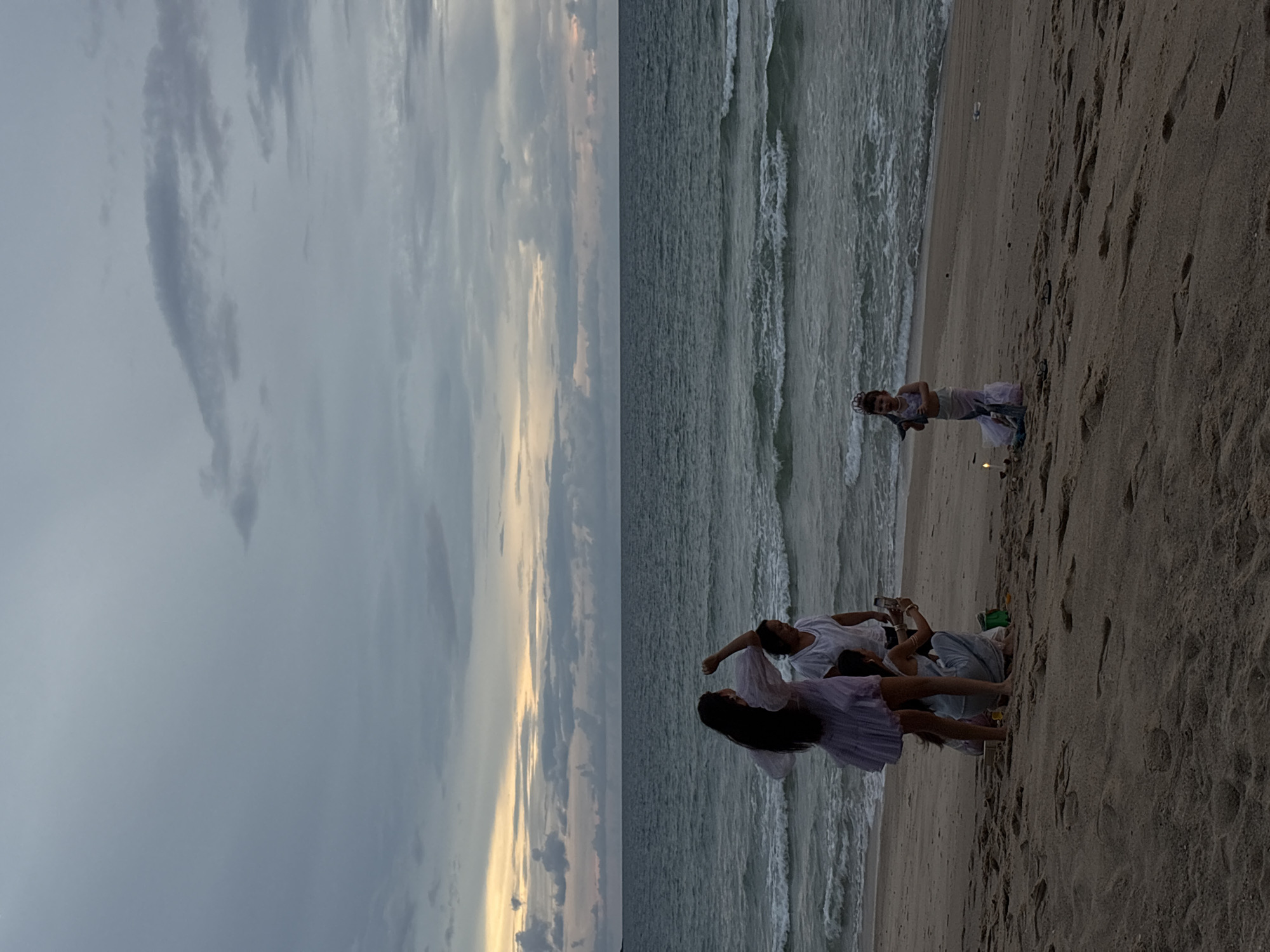 The image depicts a group of people on a sandy beach in Choeng Thale, Thailand, at dusk. The sky is overcast with dramatic clouds, and the ocean is visible with gentle waves. In the foreground, on the sand, are four individuals. A woman in a light-colored dress is standing, with her arms raised. Next to her, a person in a white t-shirt is looking at a smartphone. Another person is partially visible below them, also seemingly engaged with a phone. Further to the right, a young child dressed in a mermaid costume, complete with a tail and a tiara, stands near a small, lit candle. The overall scene suggests a casual gathering on the beach as the day concludes.