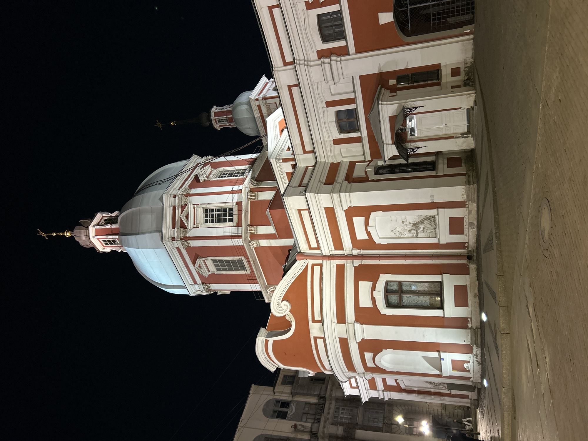 This is a nighttime shot of the St. Andrew’s Cathedral in Saint Petersburg, Russia. The cathedral is illuminated and displays its red and white baroque facade. The building has a dome and a tower topped with a cross. The sky is dark and clear. A street and surrounding buildings are partially visible at the bottom of the shot.