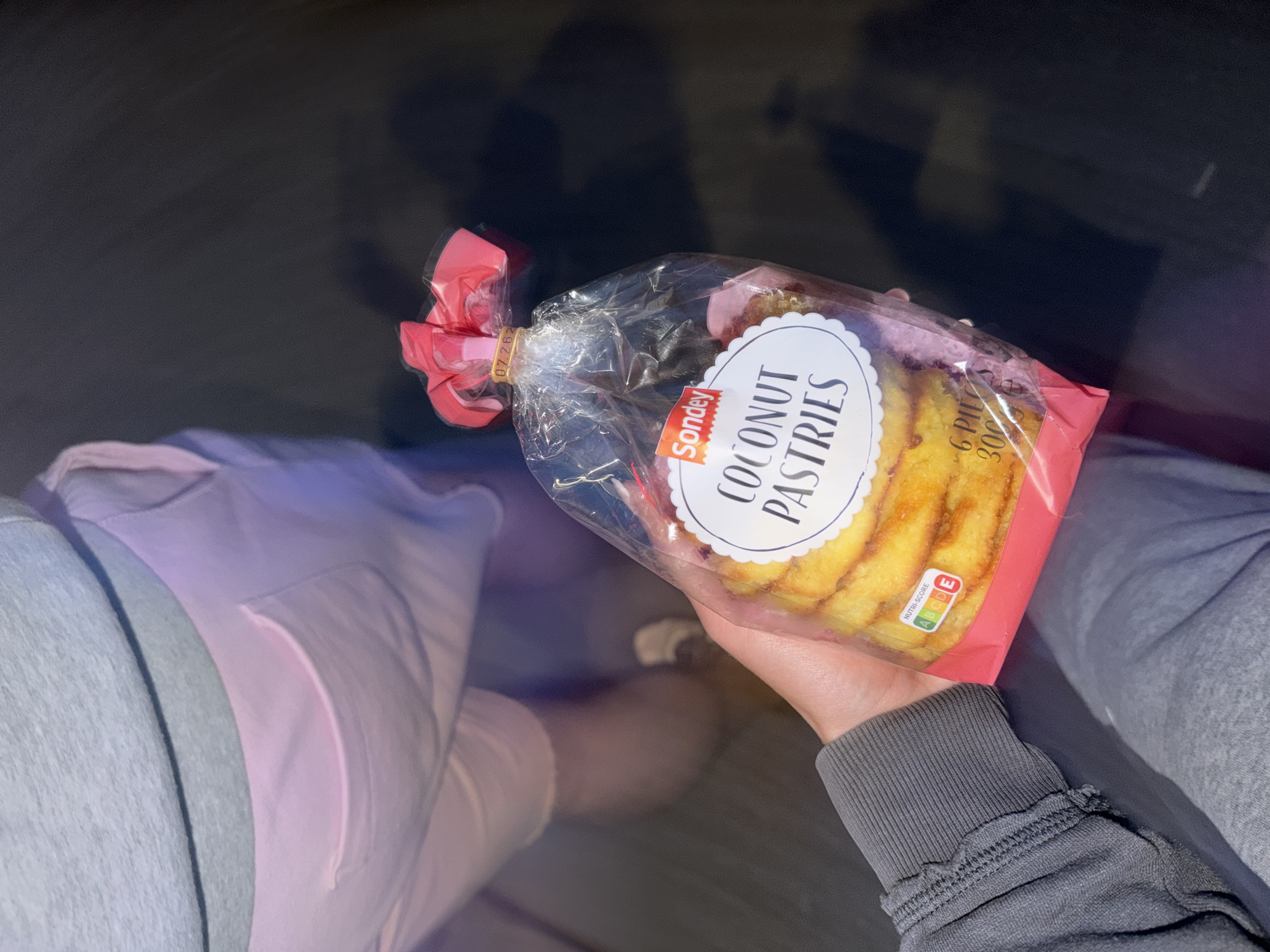 The image shows a person seated, holding a package of "Sondey Coconut Pastries". The person is wearing grey sweatpants on the right leg and light pink sweatpants with a pocket on the left leg. The person's feet and lower legs are visible beneath the pants. The package of pastries is sealed in clear plastic with a pink section. The background is dark and slightly blurred. There is no indication that this image was taken in Sofia, Bulgaria.