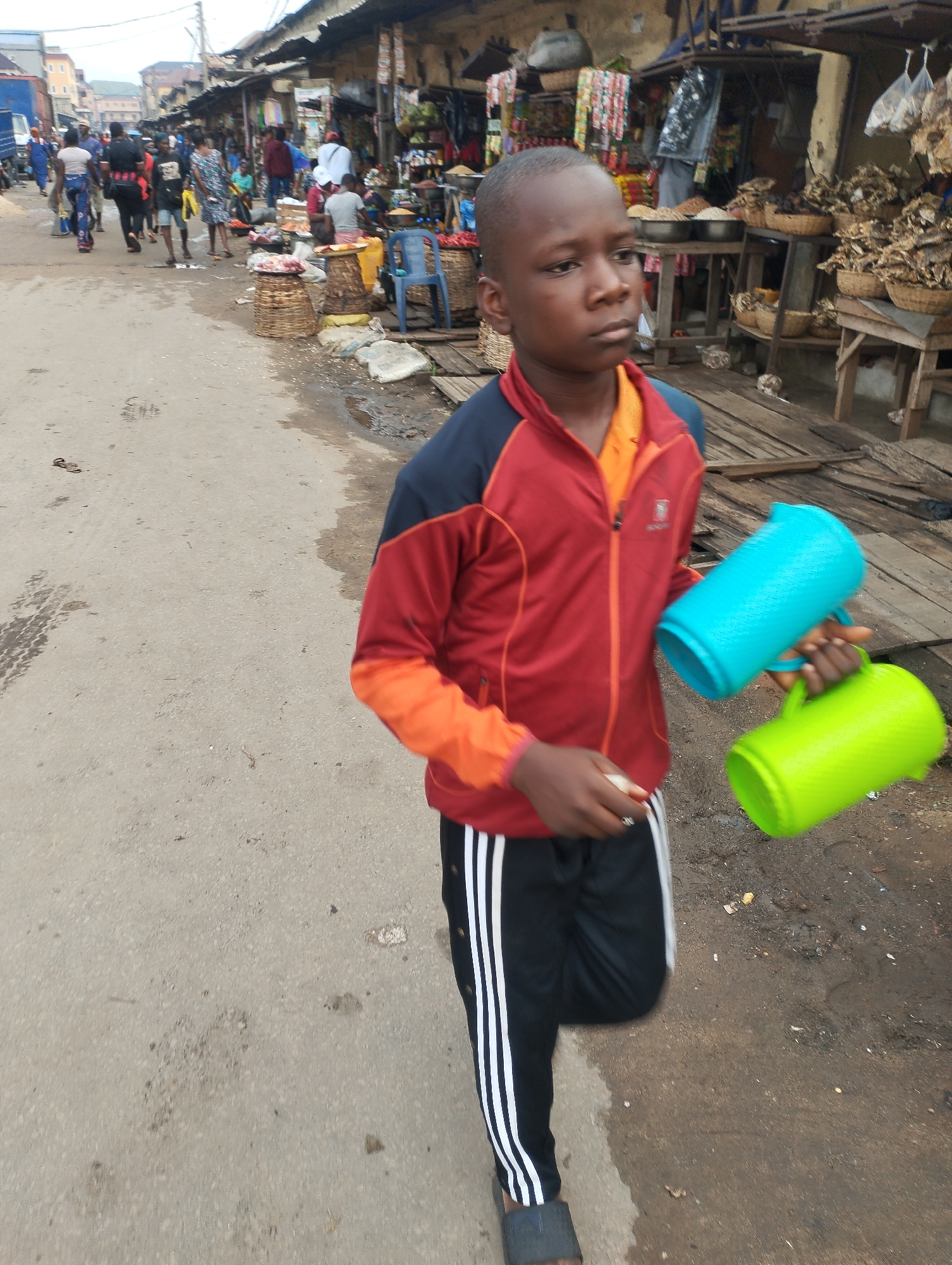 A boy is walking in a market setting, possibly in Fegge, Nigeria. He is carrying a blue plastic pitcher in his right hand and a green one in his left. He is wearing a red, orange, and blue jacket with black pants and sandals. The street is paved, and there are market stalls and people in the background. There are signs of commerce and pedestrian traffic.